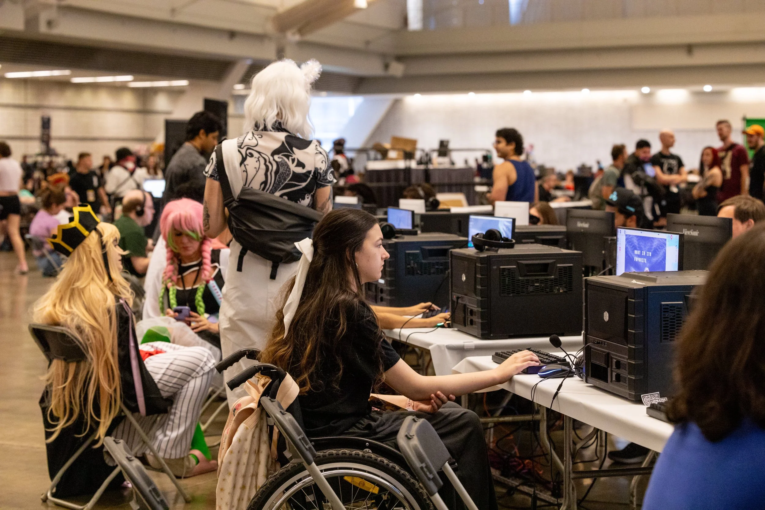 People participating in a gaming event at a large indoor venue, with some wearing cosplay costumes and others gaming on desktop computers.