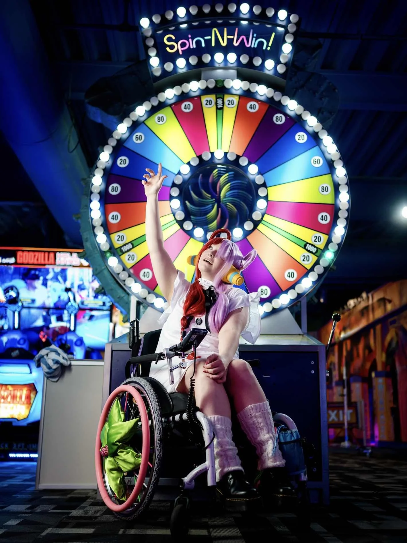 A person in a wheelchair with colorful pink and purple hair, dressed in a costume, sitting in front of a large spinning wheel game at an indoor arcade or amusement park, illuminated by bright lights.