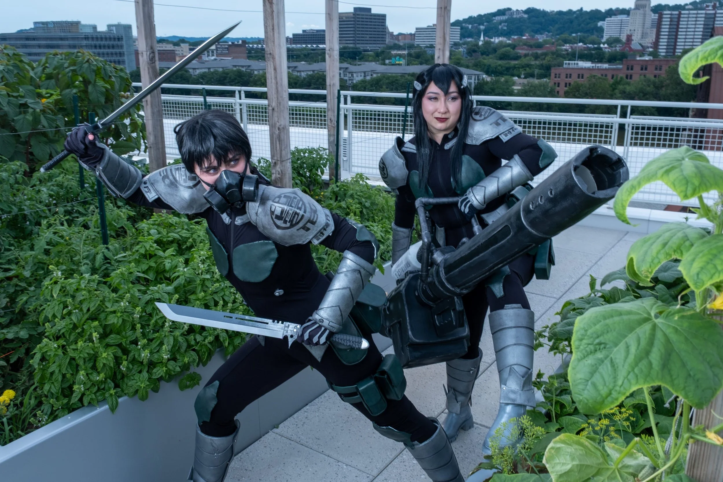 Two women dressed in futuristic costumes with armor and weapons on a rooftop garden, with city buildings and a hill in the background.
