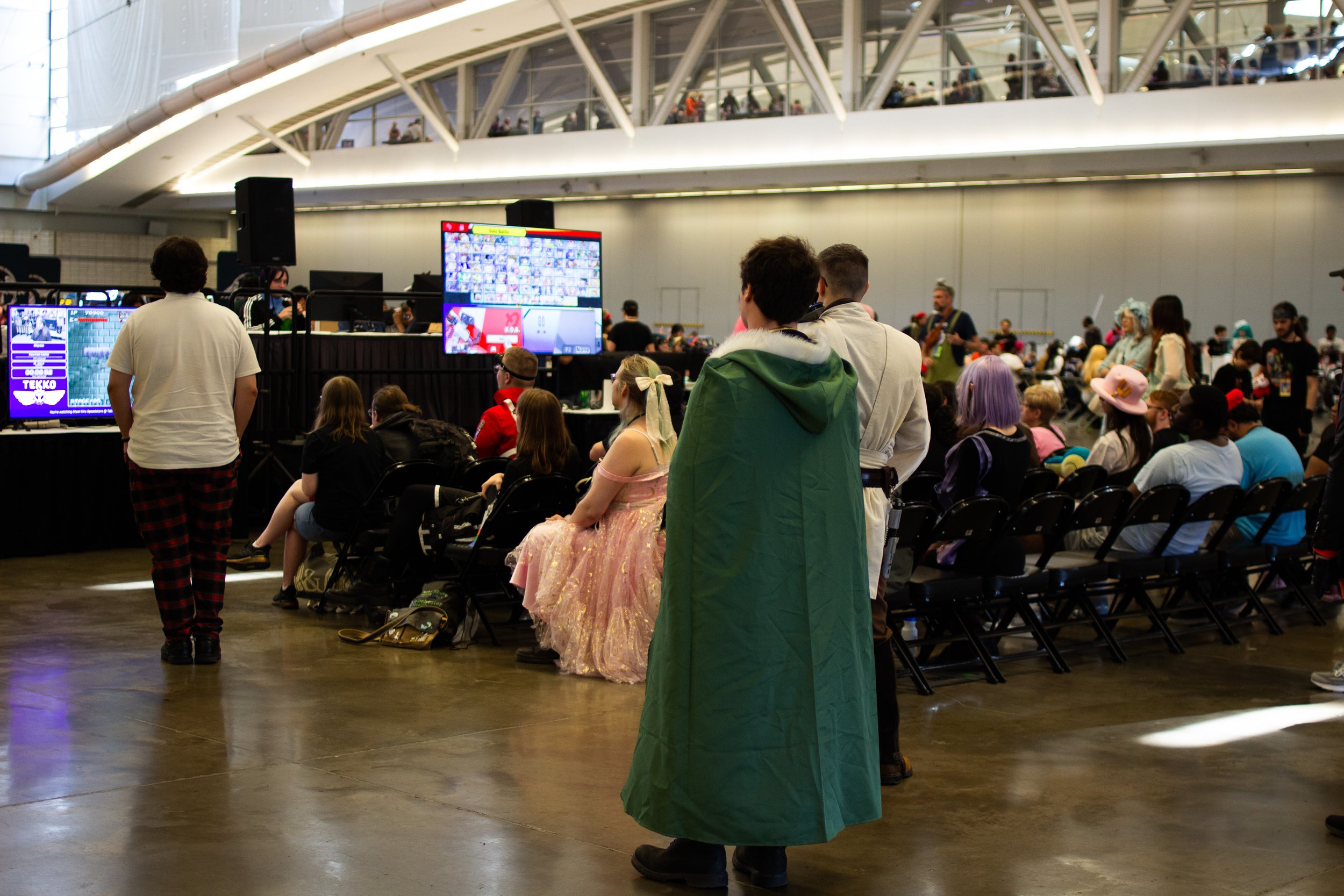 People sitting and standing at an indoor event, some watching a large screen, with others walking around, in a spacious, modern venue with multiple levels.