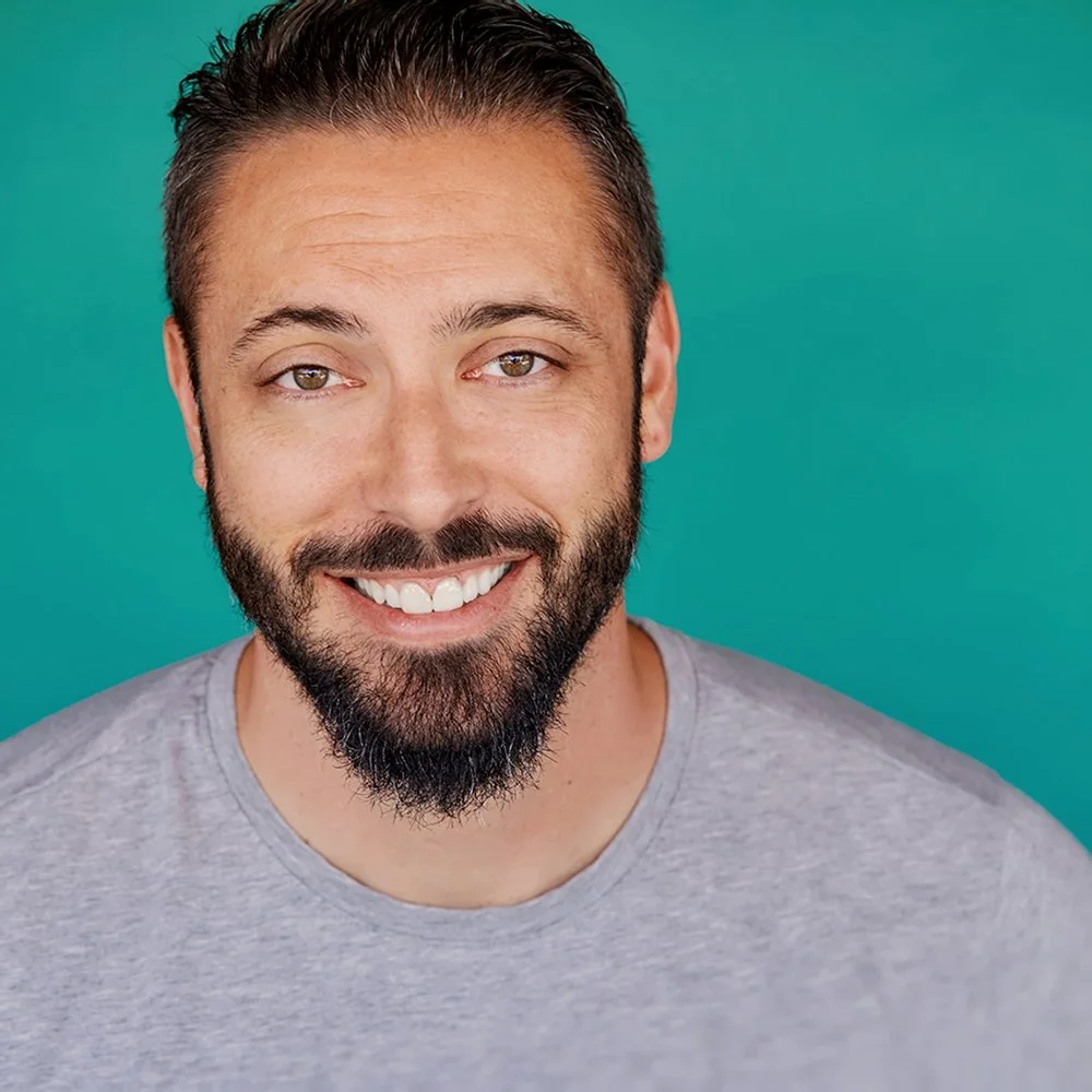 A smiling man with a beard and mustache, wearing a light gray t-shirt, poses against a teal background.