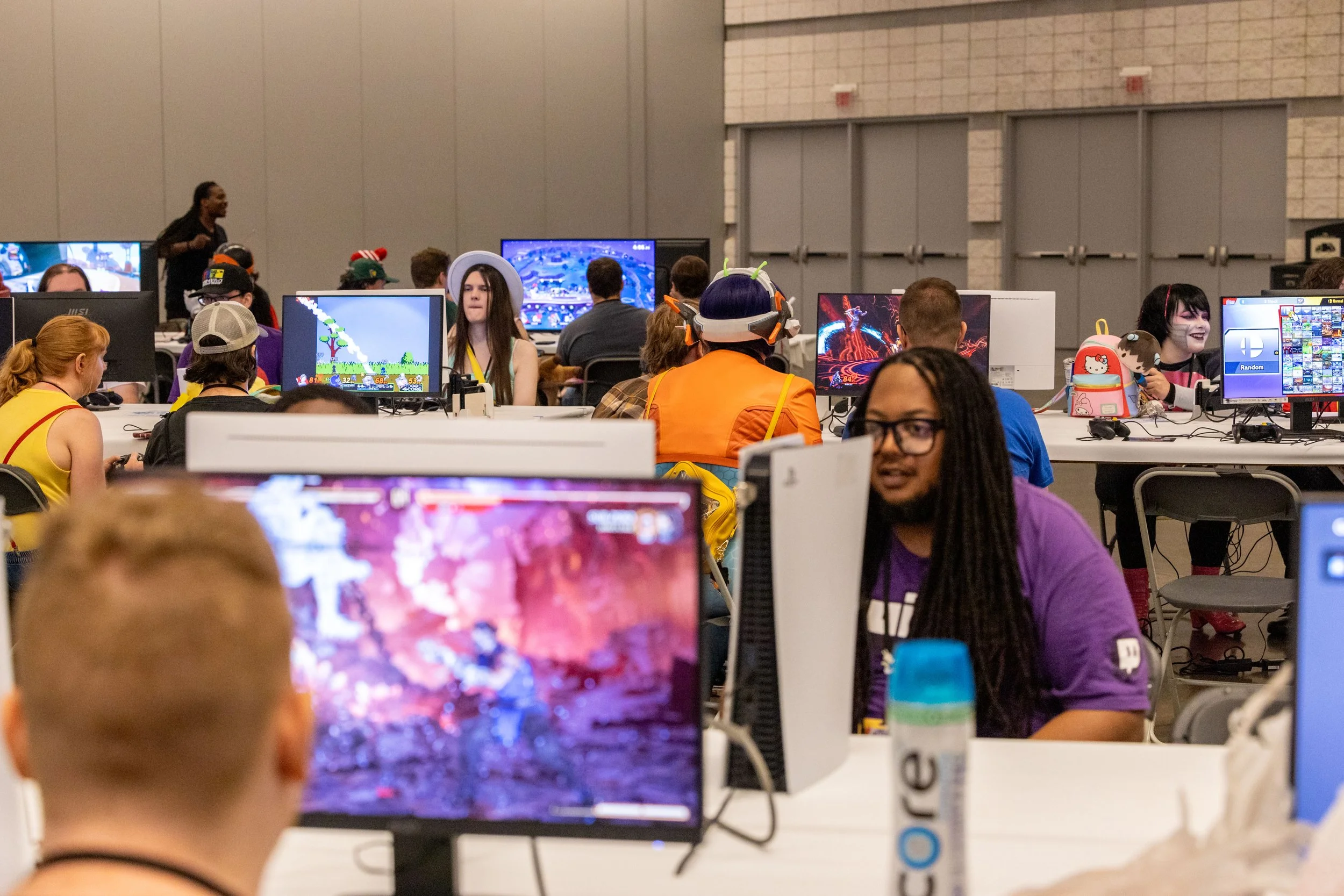 People gathered at a gaming event playing on desktop computers with colorful screens, some wearing costumes or hats, in a large indoor space with gray walls and multiple monitors visible.