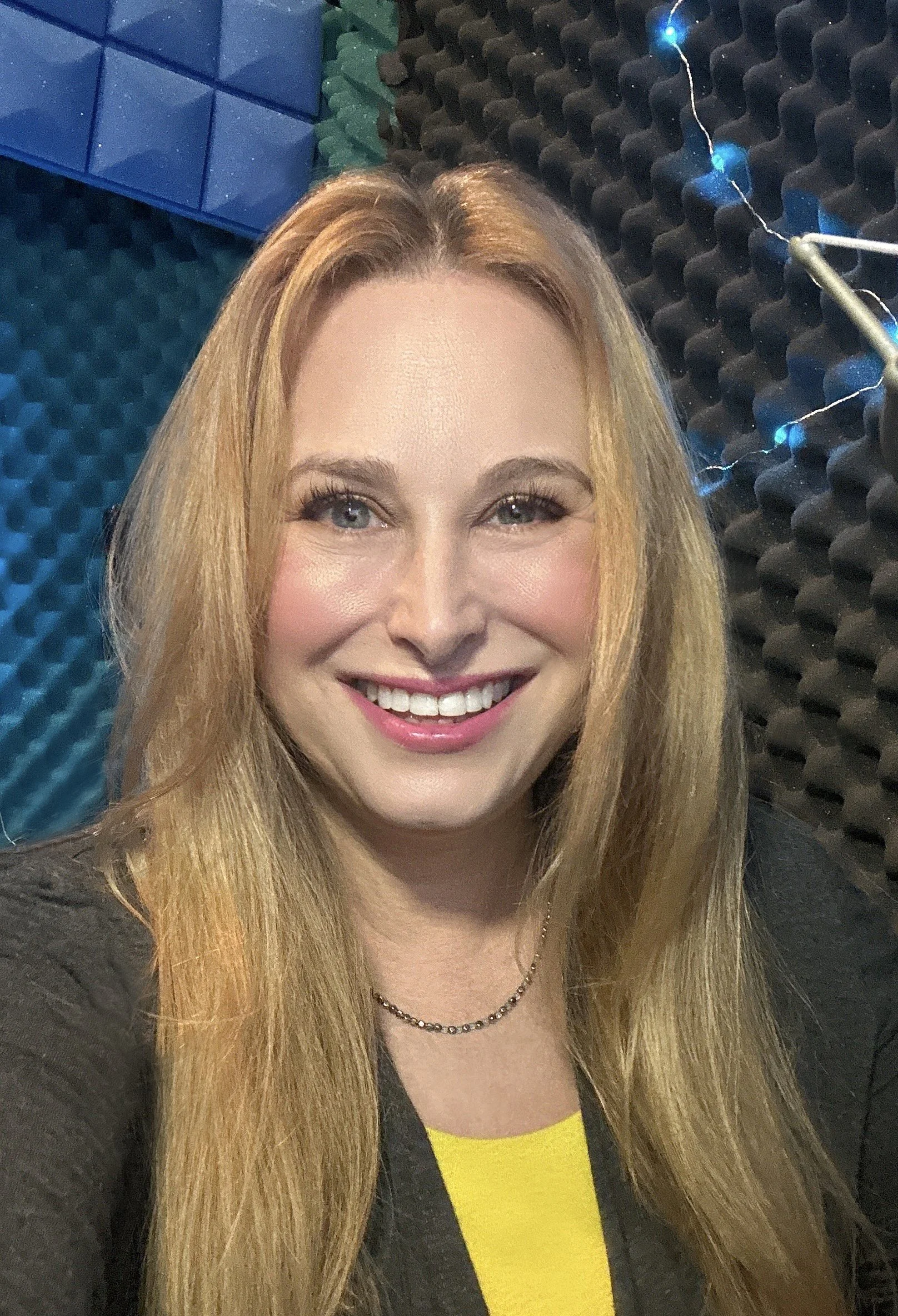 Close-up of a woman with long blonde hair smiling, wearing a black top with a yellow shirt underneath, and a black necklace. The background features soundproof foam panels.