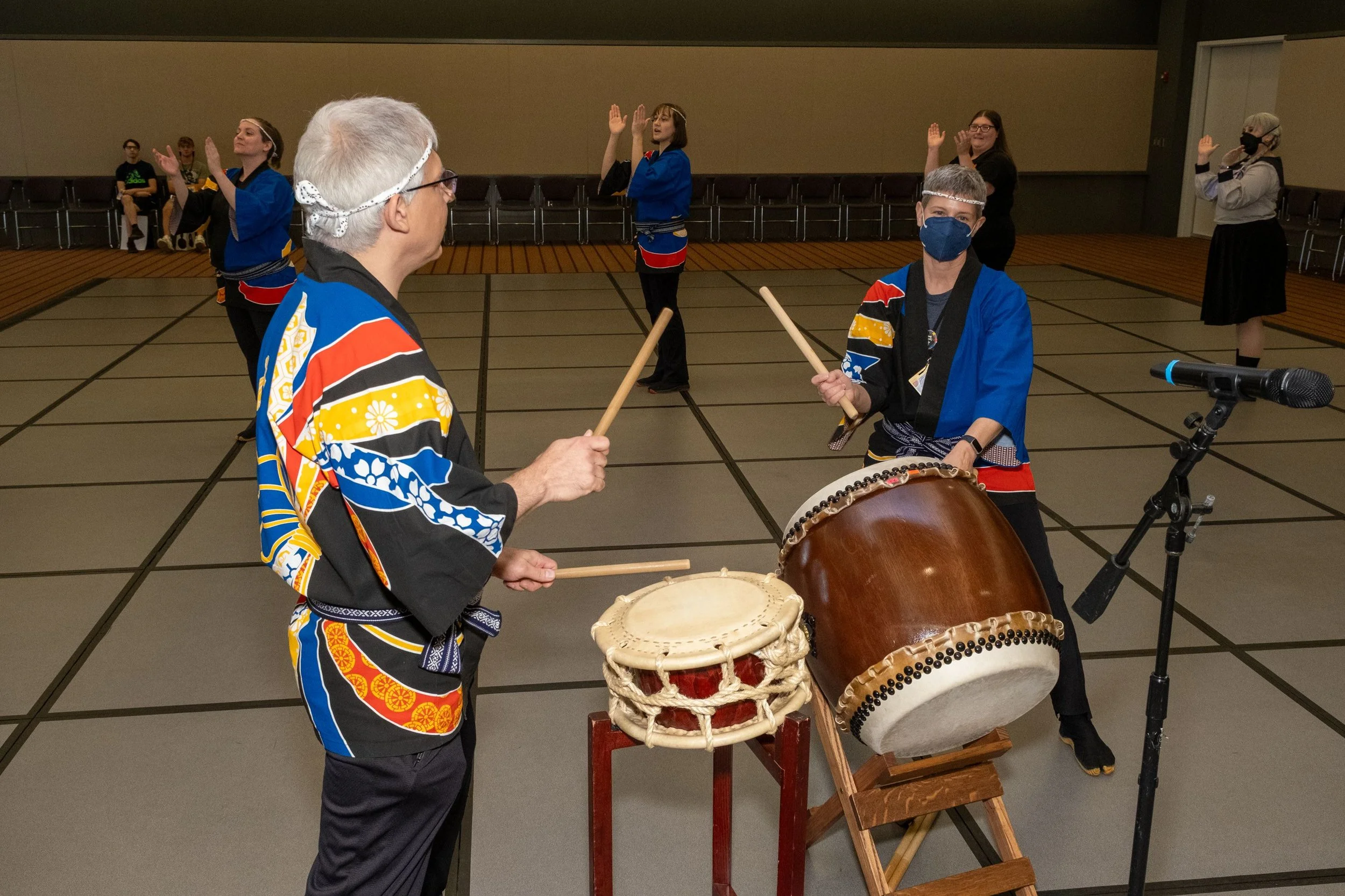 Multiple people wearing masks and colorful traditional clothing, playing drums in an indoor space with empty chairs along the walls.