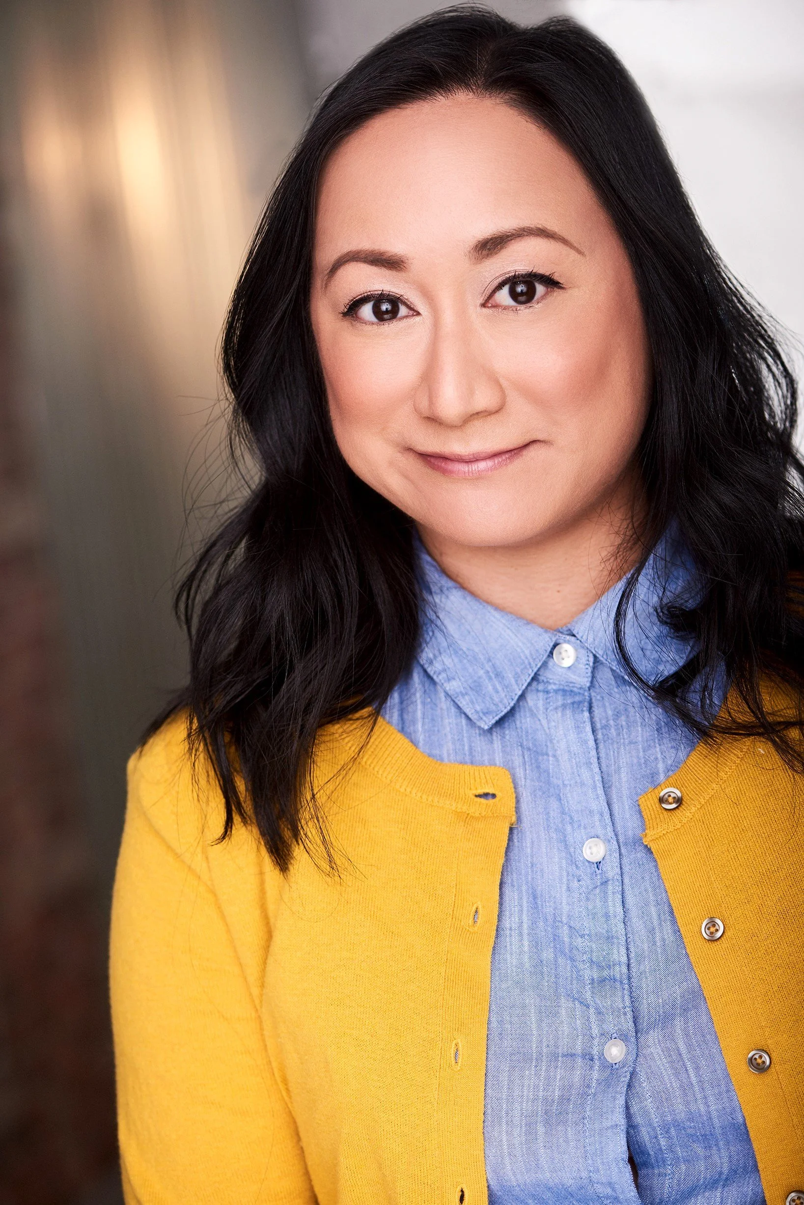 A woman with long black hair and a light complexion smiling, wearing a blue collared shirt and a yellow cardigan.