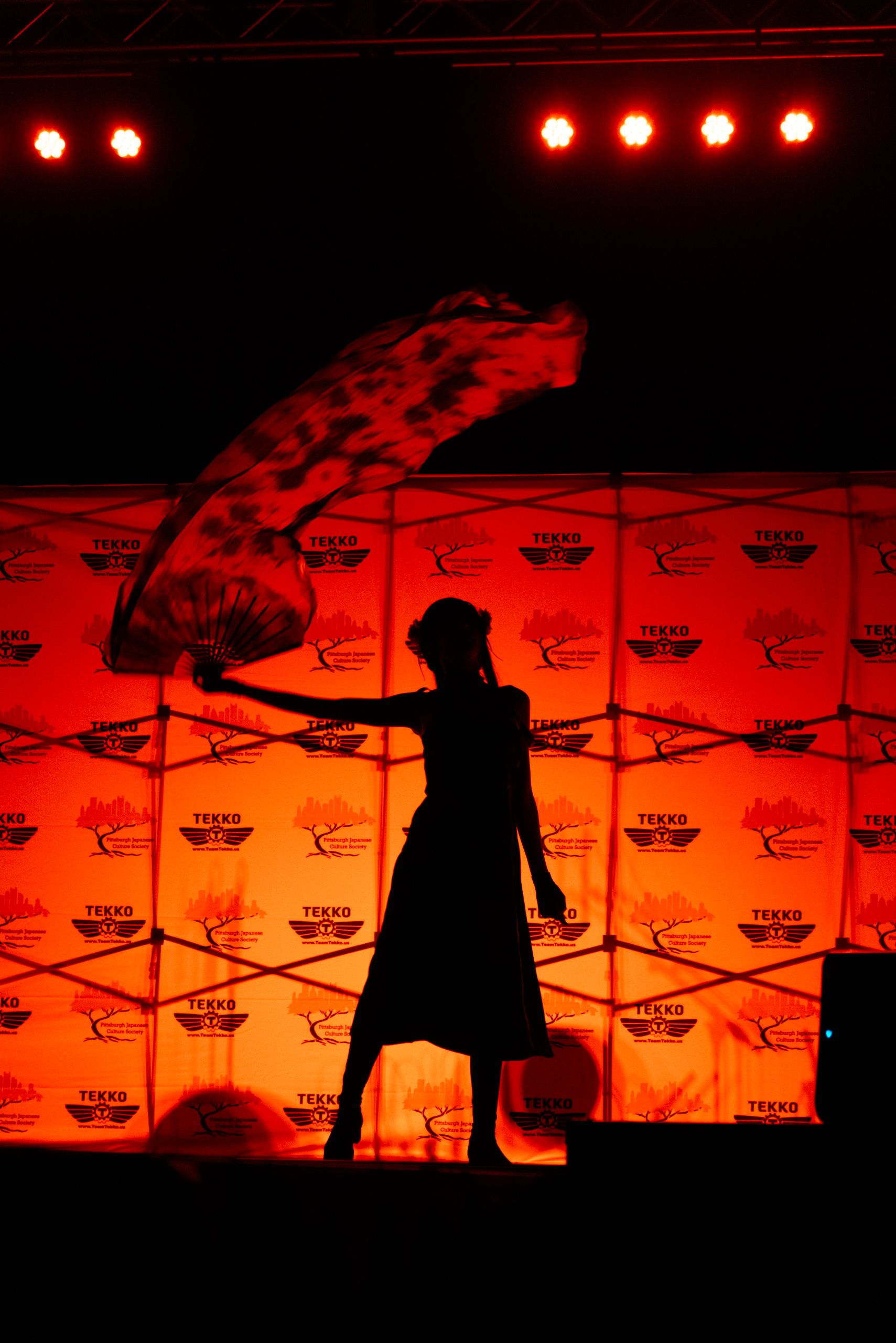A backlit cosplayer waves fans and veils on stage during a performance.