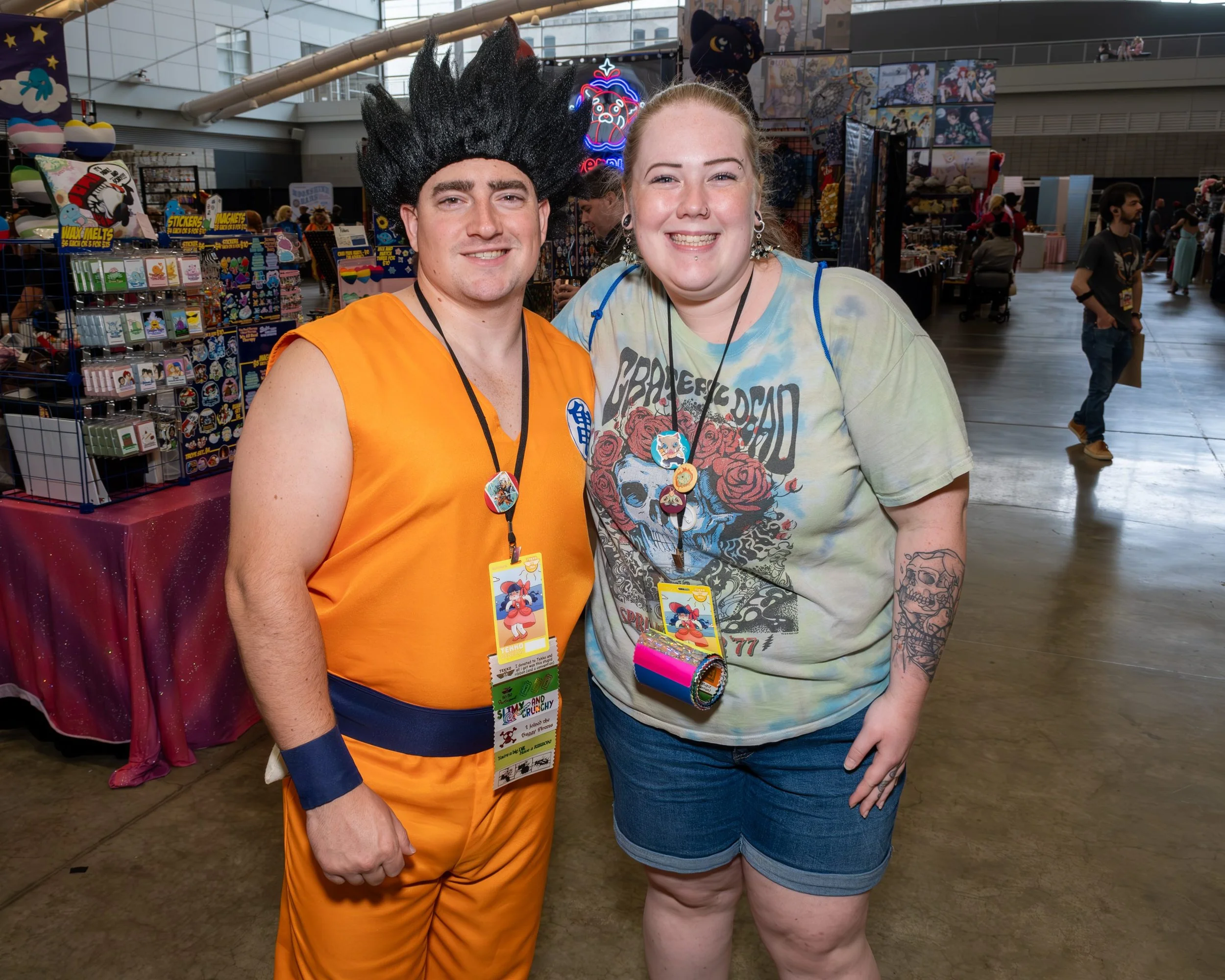 Two people standing close together at a convention or event, smiling at the camera. The person on the left is dressed as Goku in an orange costume with black spiky wig, and the person on the right has long blonde hair, tattoos on her arm, and is wear