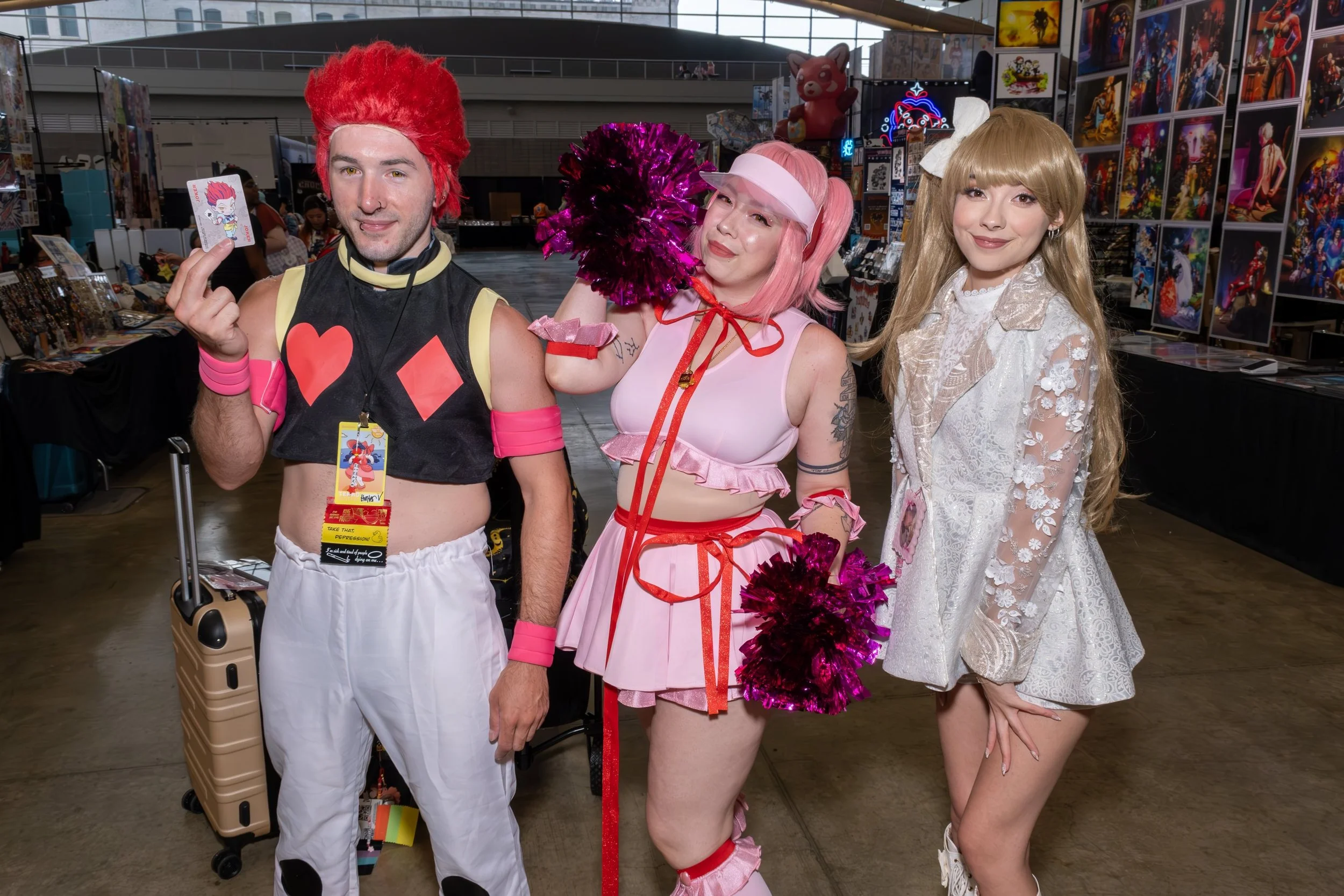 Three people in colorful costumes at a convention. The person on the left is dressed in a black and pink outfit with a large red wig, holding a card and a small suitcase. The person in the middle is dressed as a pink-haired anime character with pom-p