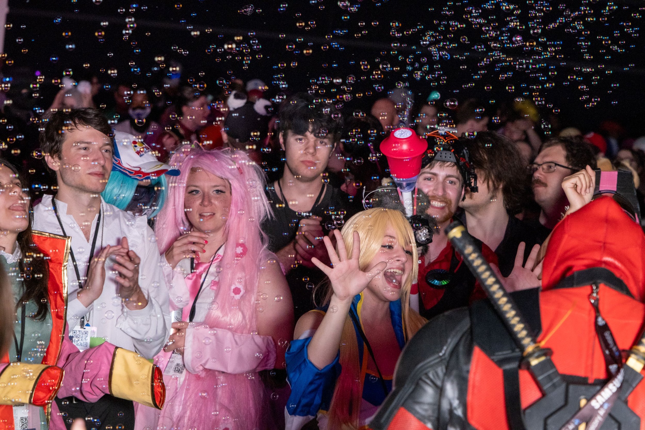 A group of people at a lively event, with some dressed in colorful costumes and others in casual attire, surrounded by floating bubbles.