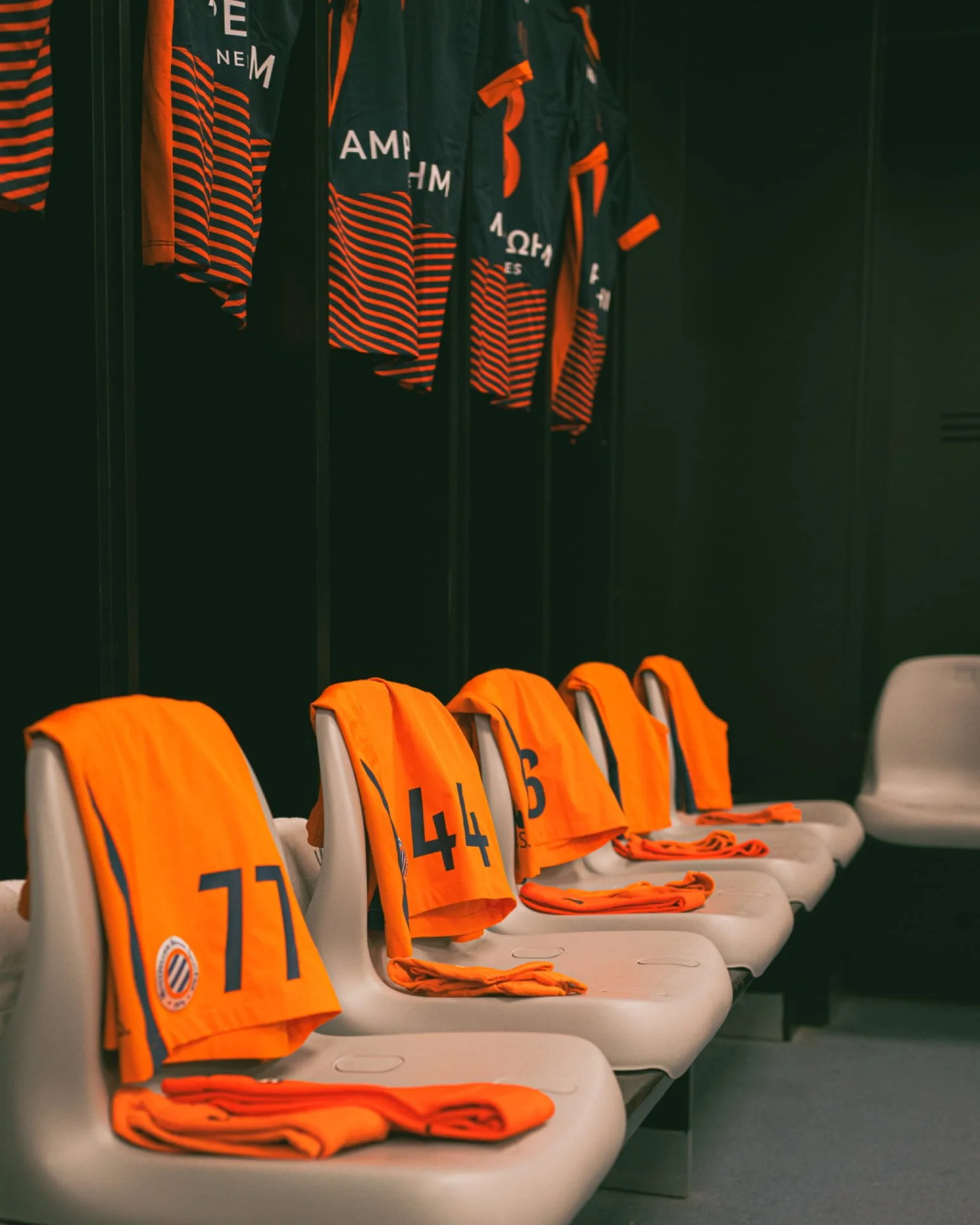 Des maillots de sport orange avec des numéros et un drapeau au dos, suspendus dans un vestiaire, avec des vestes et des équipements en arrière-plan.