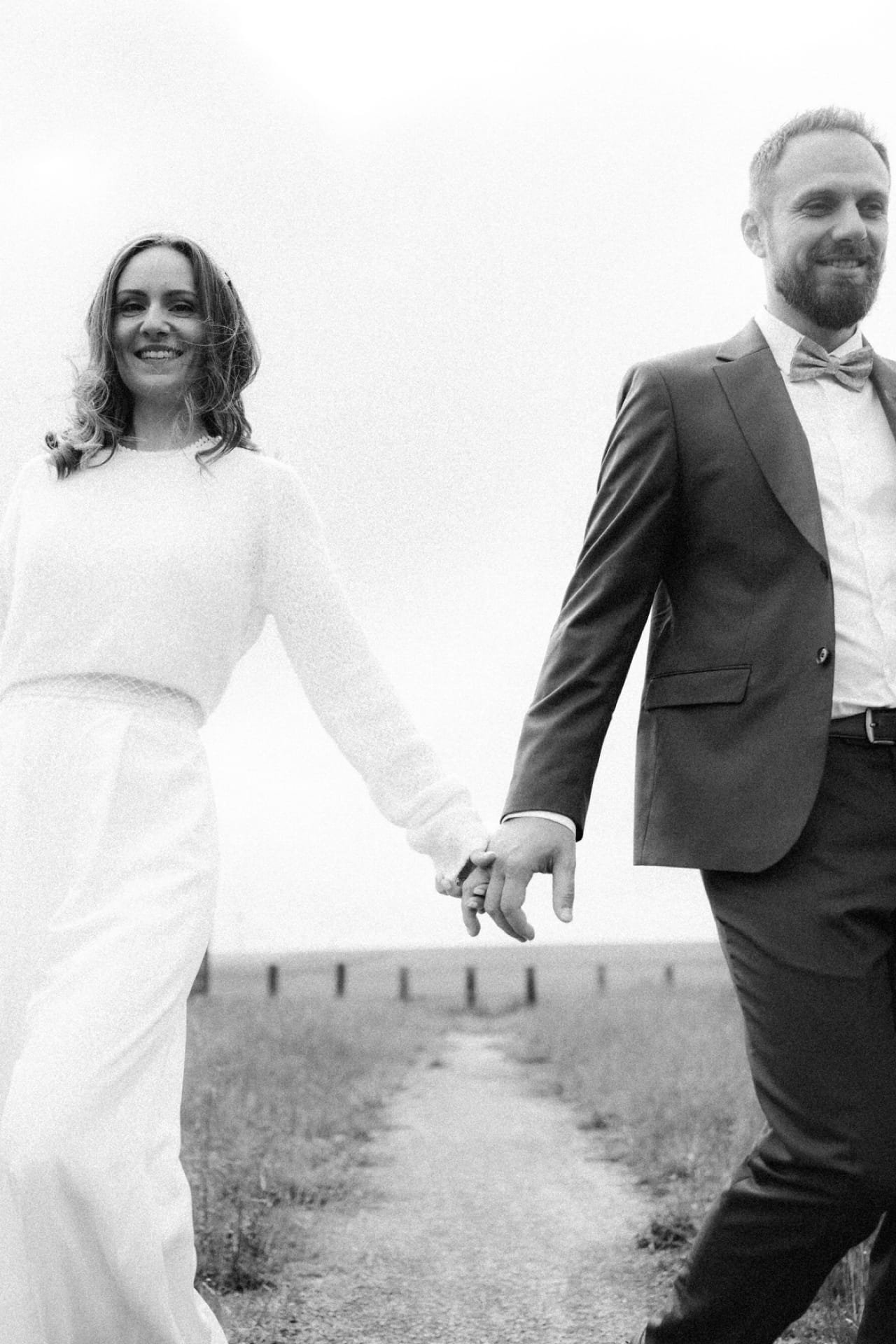 Un couple habillé élégamment, probablement lors d'un mariage, tenant la main dans un endroit en extérieur, avec une barrière en bois en arrière-plan, souriant, en noir et blanc.