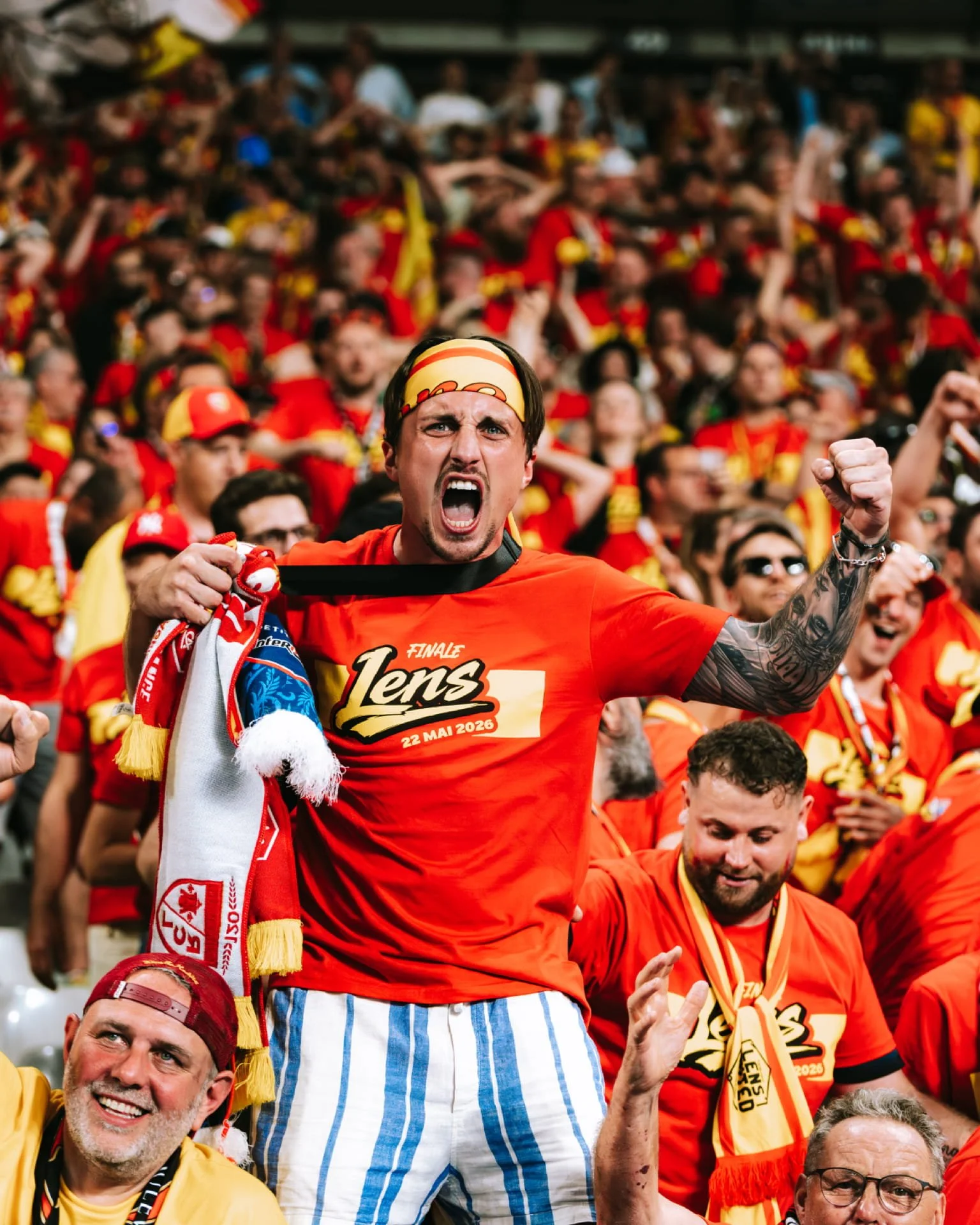 Un groupe de supporters de football, principalement en rouge, célébrant un événement sportif, avec un homme au premier plan criant et levant le poing, portant un bandeau et un t-shirt avec la date 22 mai 2026.