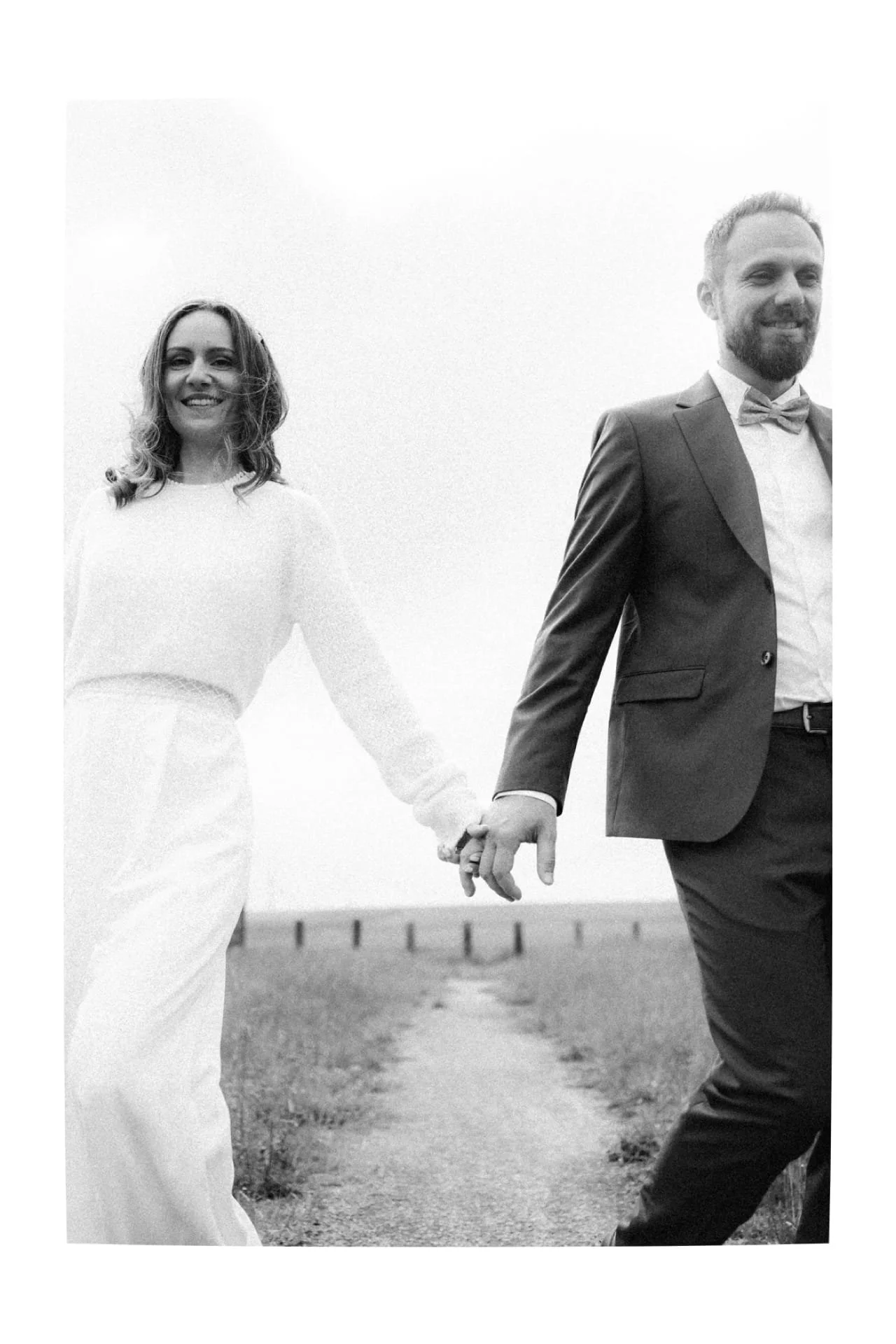 Un couple marchant main dans la main, vêtus pour une occasion spéciale, probablement un mariage, dans un décor extérieur.