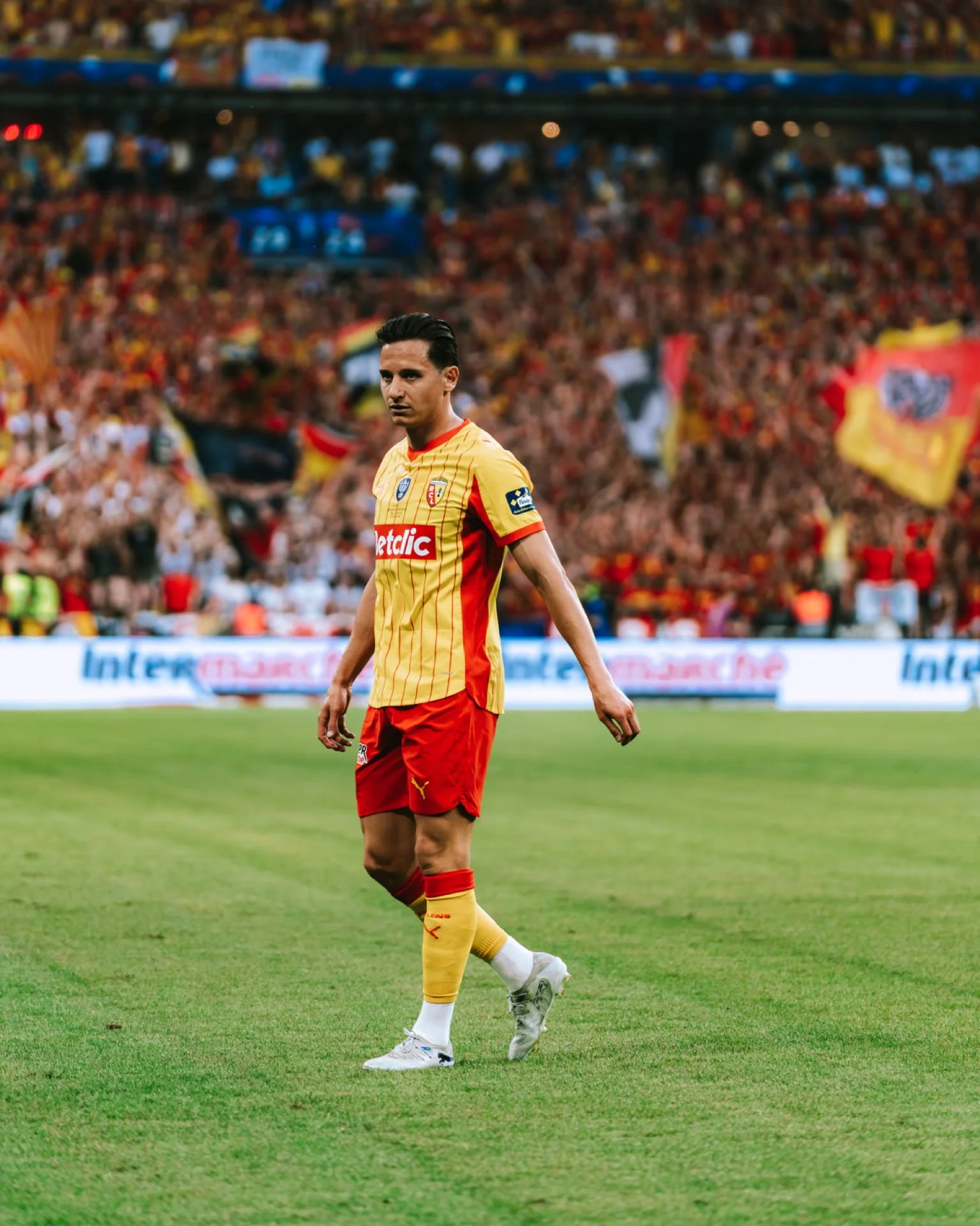 Un joueur de football dans un stade, portant un maillot jaune et rouge avec le logo de l'équipe, marche sur le terrain en regardant vers l'avant, avec un public nombreux en arrière-plan.