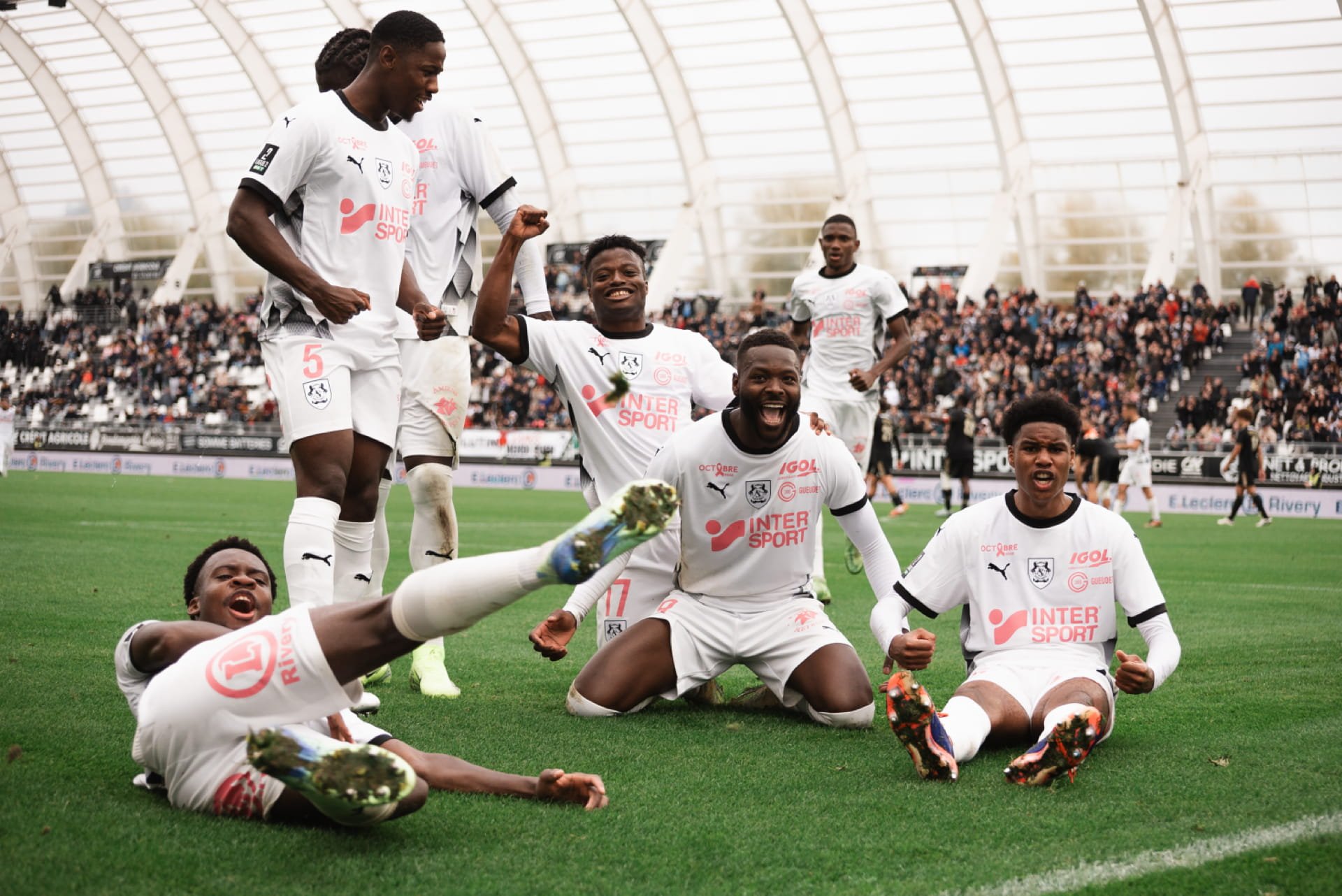 Groupe de joueurs de football en uniforme blanc, posant pour la photo dans un stade avec une foule en arrière-plan, célébrant une victoire. Cauê Foto, photographe Amiens, photographe de mariage et événementiel