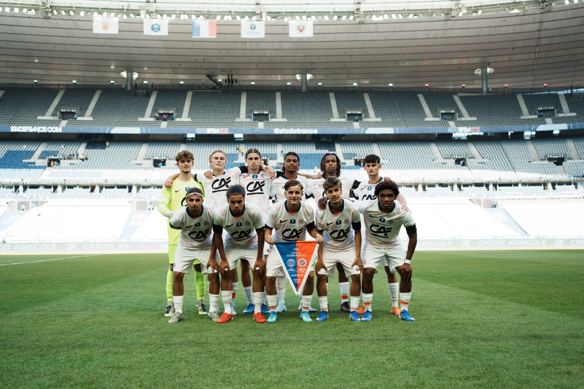 Une équipe de football posant pour une photo dans un stade vide, avec des drapeaux suspendus au-dessus.