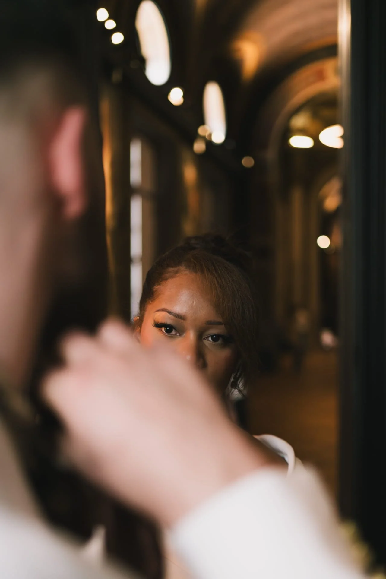 Une femme regarde son reflet dans un miroir dans un environnement chaleureux, probablement un salon ou un espace de beauté, avec une lumière douce et des arches en arrière-plan. Cauê Foto, photographe Amiens, photographe de mariage et événementiel