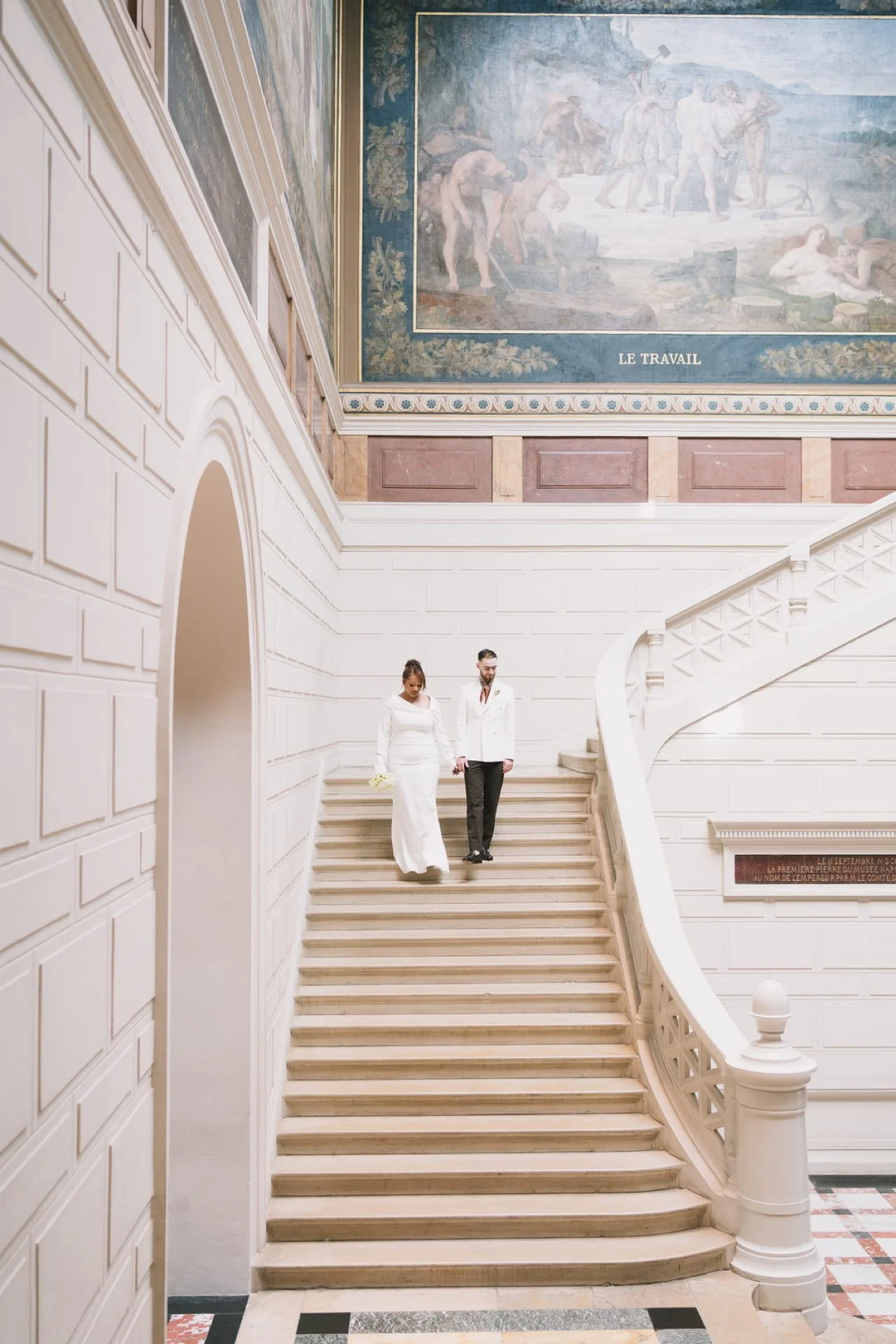 Un couple en vêtements de mariage descend un escalier en pierre dans un bâtiment élégant avec des murs blancs, une grande fresque en toile sur le mur du haut, et un sol en carreaux multicolores. Cauê Foto, photographe à Amiens 