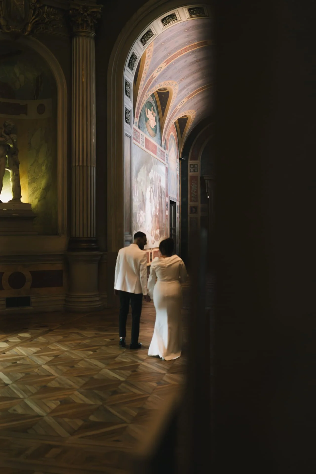 Deux personnes, un homme et une femme habillés en blanc, marchent dans une salle ornée de fresques et de sculptures, avec un sol en parquet en motif géométrique. Cauê Foto, photographe Amiens, photographe de mariage et événementiel