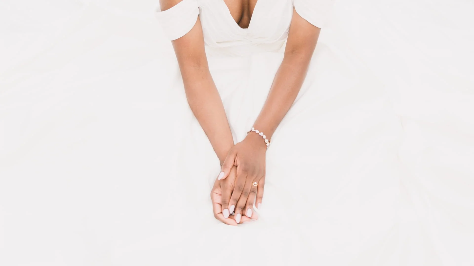 Mains féminines avec manucure blanche, portant bracelets et bague, positionnées sur un fond blanc, probablement en position de prière ou d'attente.