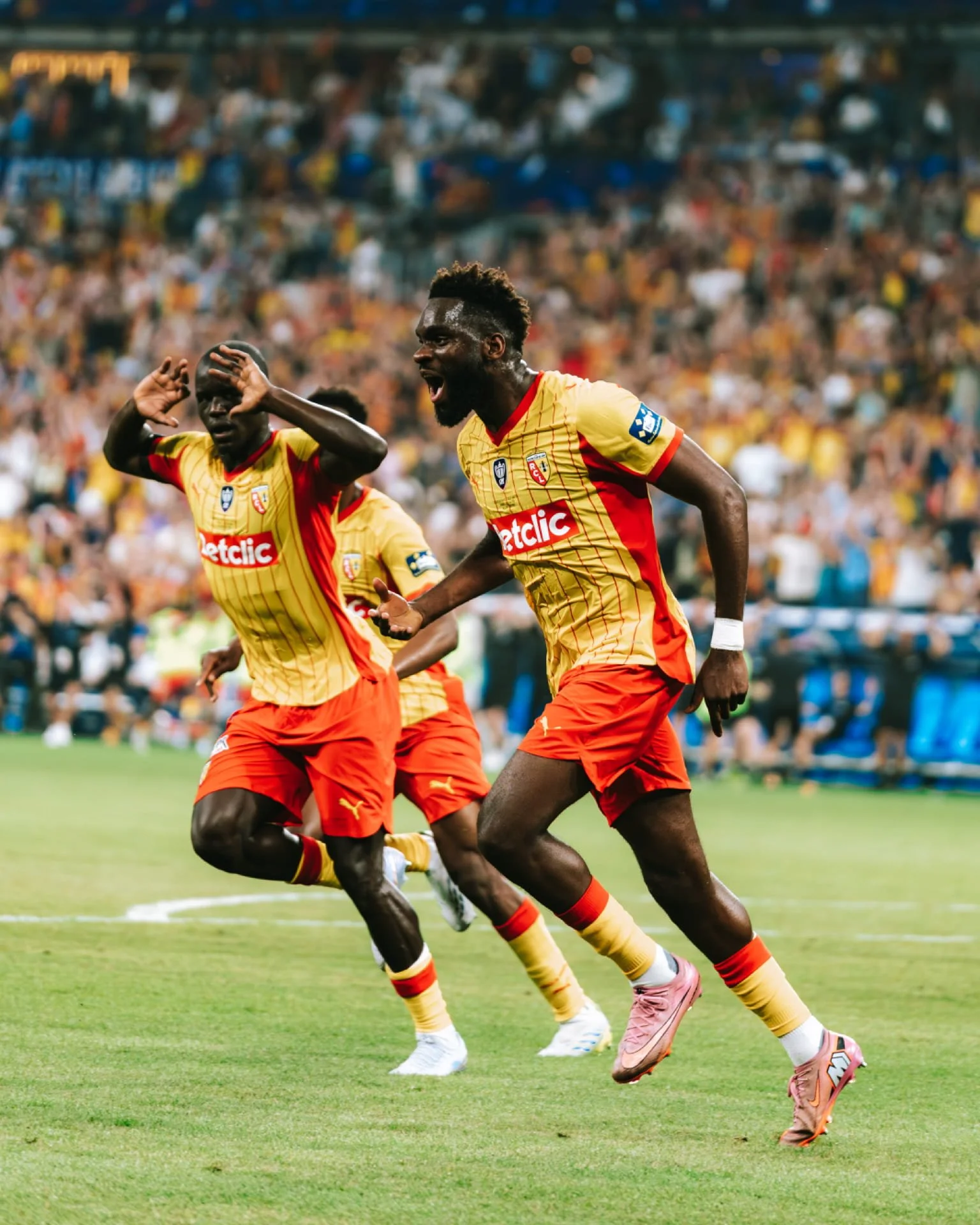 Deux joueurs de football en action sur le terrain, portant des maillots jaunes et rouges, avec un public nombreux en arrière-plan.