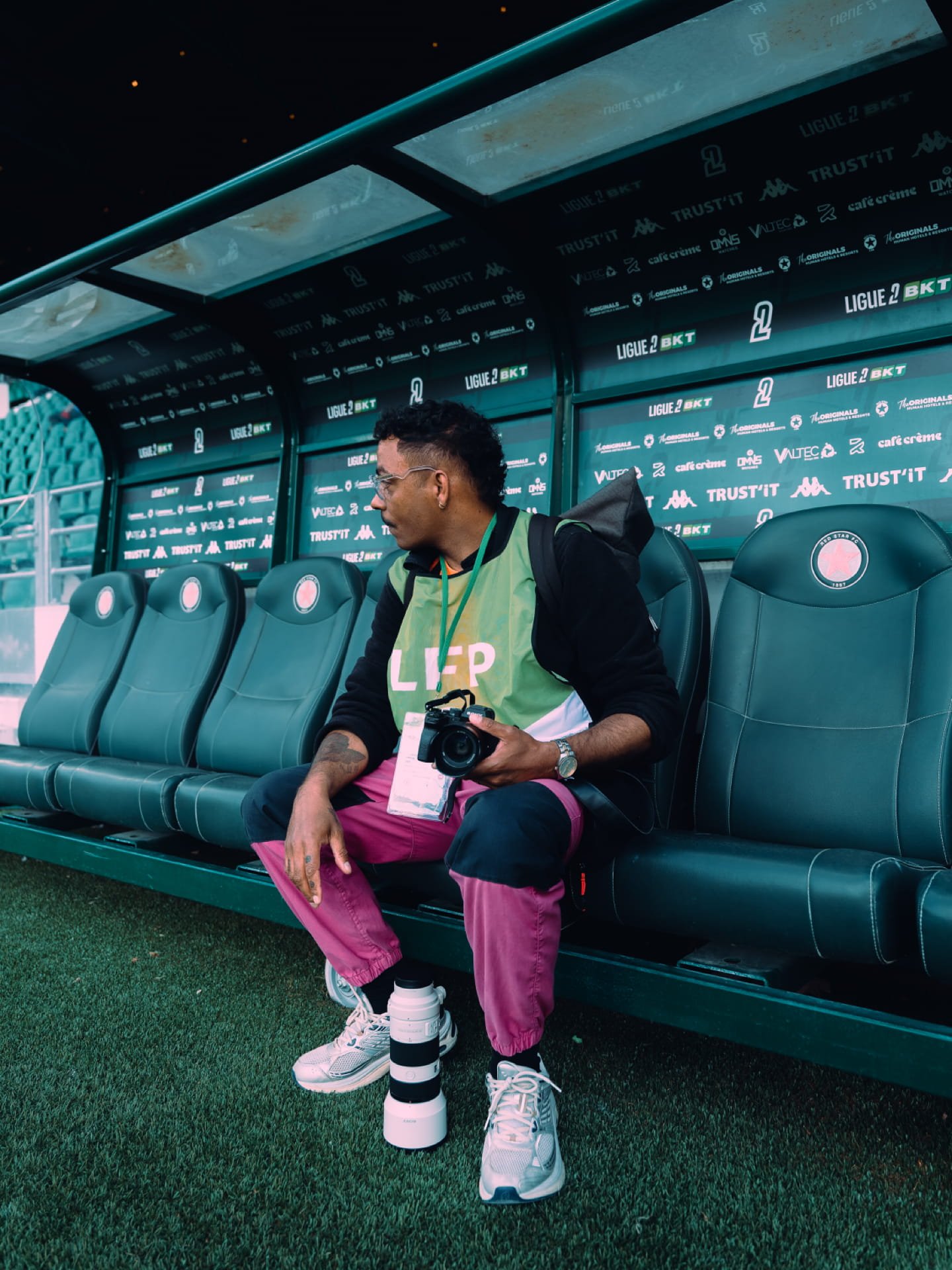 Un homme assis sur un banc de touche d'un stade, tenant un appareil photo, portant des vêtements colorés, des lunettes et des chaussures blanches, avec un gros téléobjecteur de caméra posé à ses pieds.