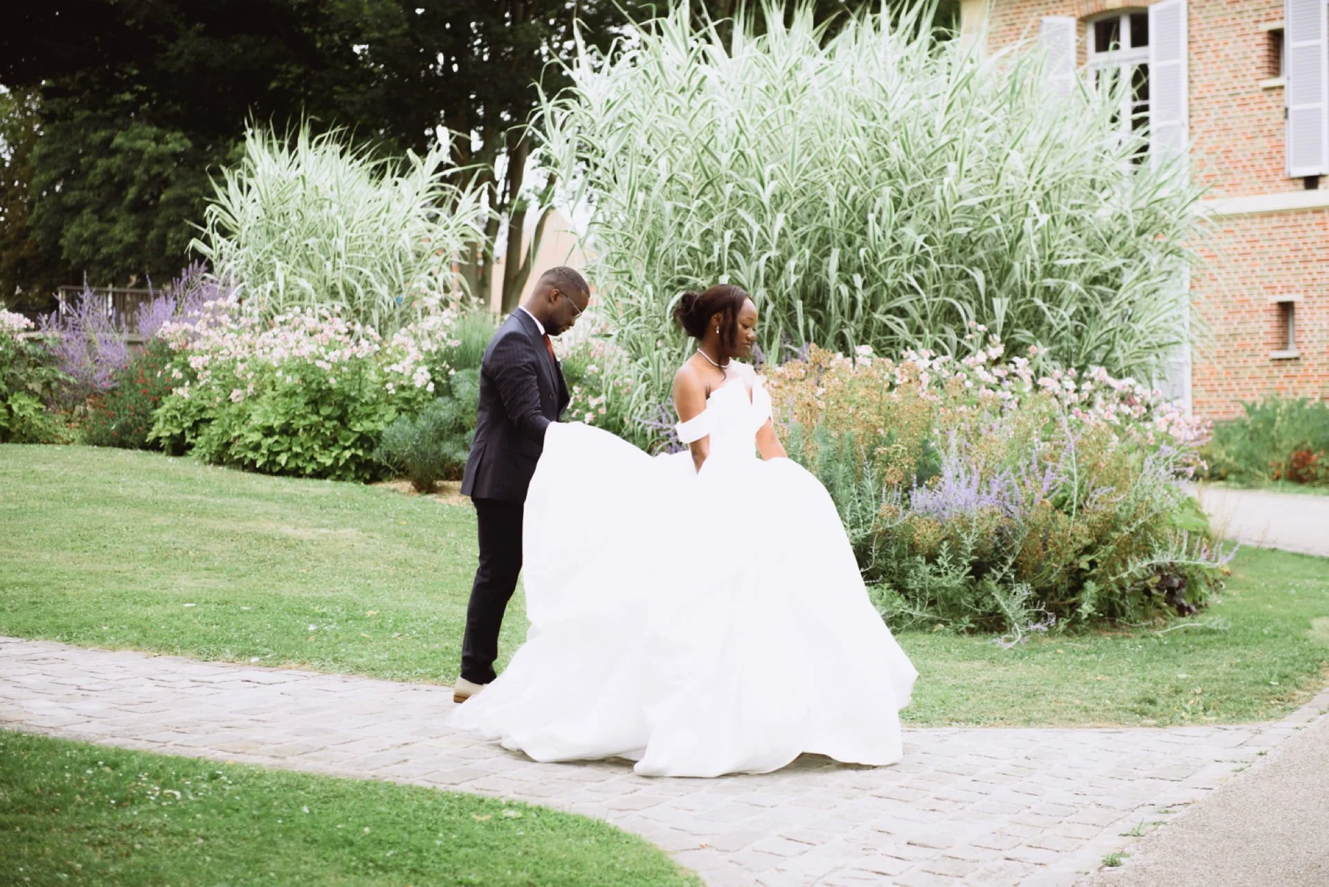 Un couple habillé en vêtements de mariage se promène dans un jardin fleuri avec un bâtiment en brique en arrière-plan.