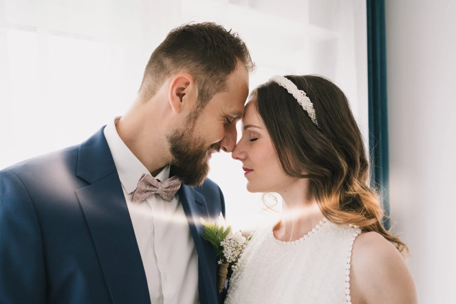 Un couple de mariage, leur front et leurs nez se touchent, ils ont l'air très heureux et tendres, la femme porte une robe blanche et un bandeau de perles, l'homme porte un costume bleu avec une chemise blanche et un nœud papillon, ils semblent s'appr