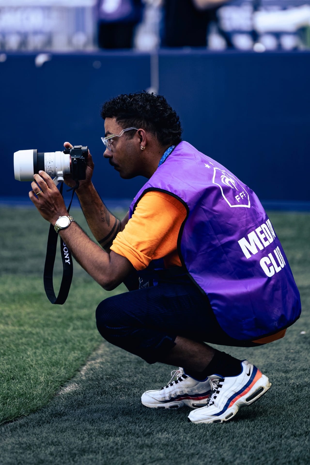 Cauê Foto, photographe de mariage , portrait et événementiel. en position accroupie, à l'extérieur sur un terrain de sport. Il porte une veste violette avec le logo de la Fédération Française de Football et des baskets blanches.