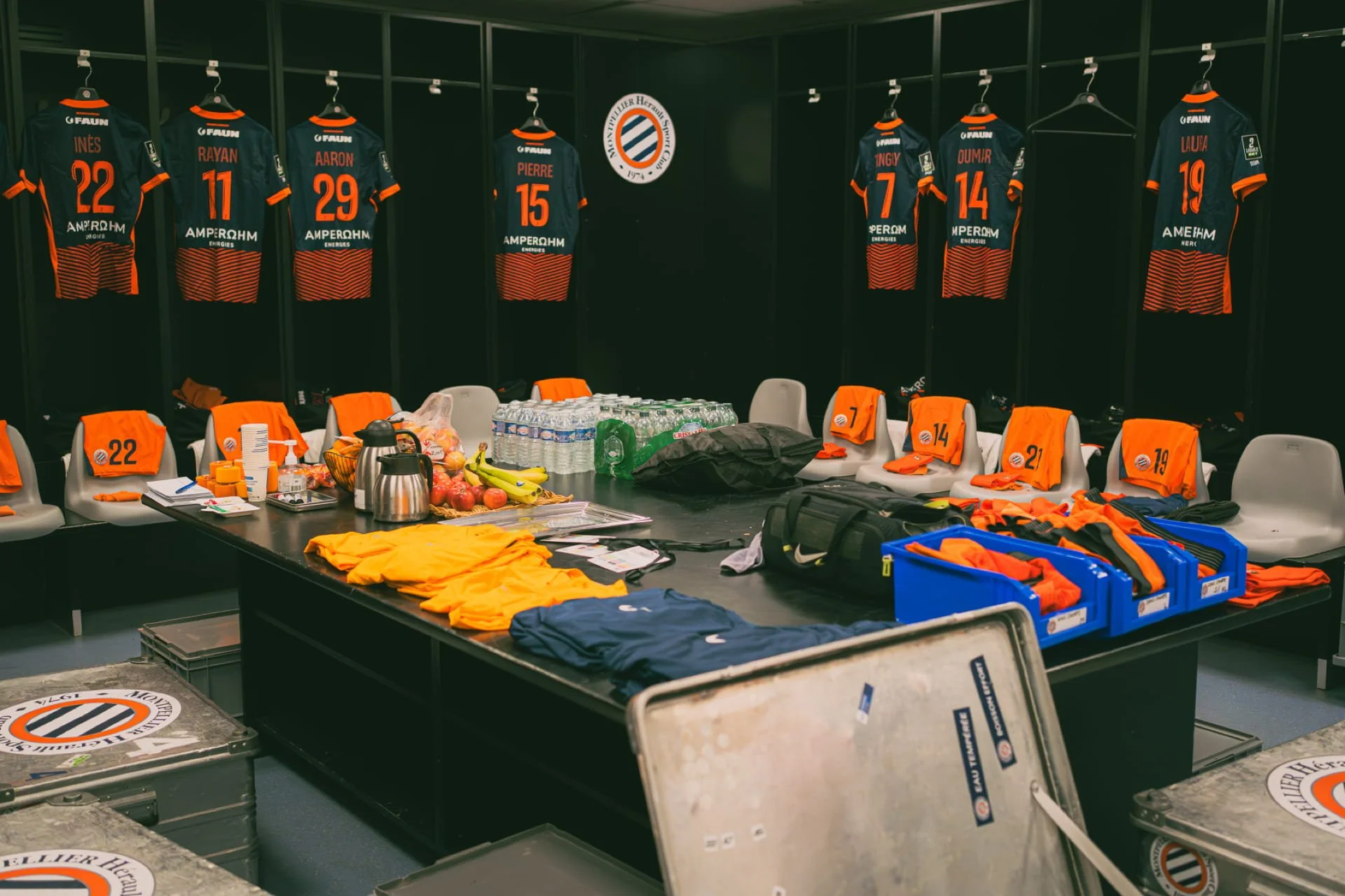 Vestiaires du club de football avec maillots suspendus portant les noms et numéros de joueurs, un tableau de verrouillage, table avec boissons, fruits et équipement sportif, logo du club sur le mur.
