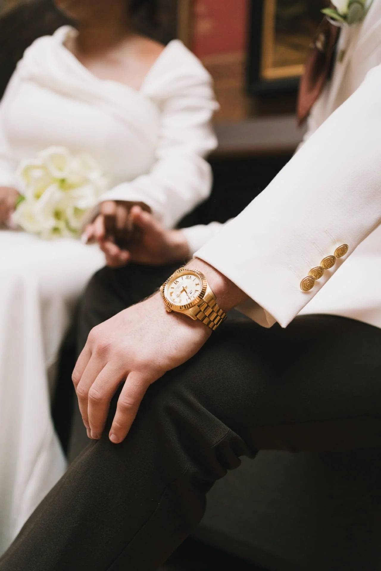 Une personne en costume blanc porte une montre en or, avec des boutons en or sur la manche de sa veste, l'autre personne porte une robe blanche et tient un bouquet de fleurs. Le cadre semble être une cérémonie ou un mariage.