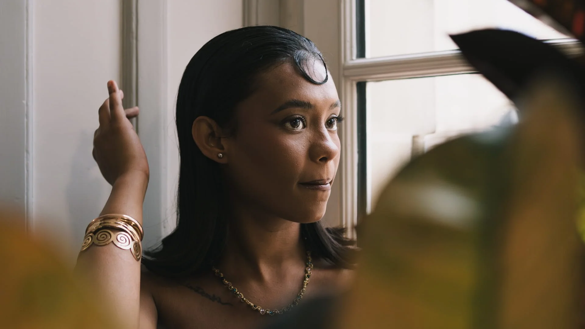 Une femme aux cheveux noirs lisses regarde par la fenêtre, avec des bijoux dorés et un tatouage visible sur le haut de son bras, dans un intérieur lumineux.