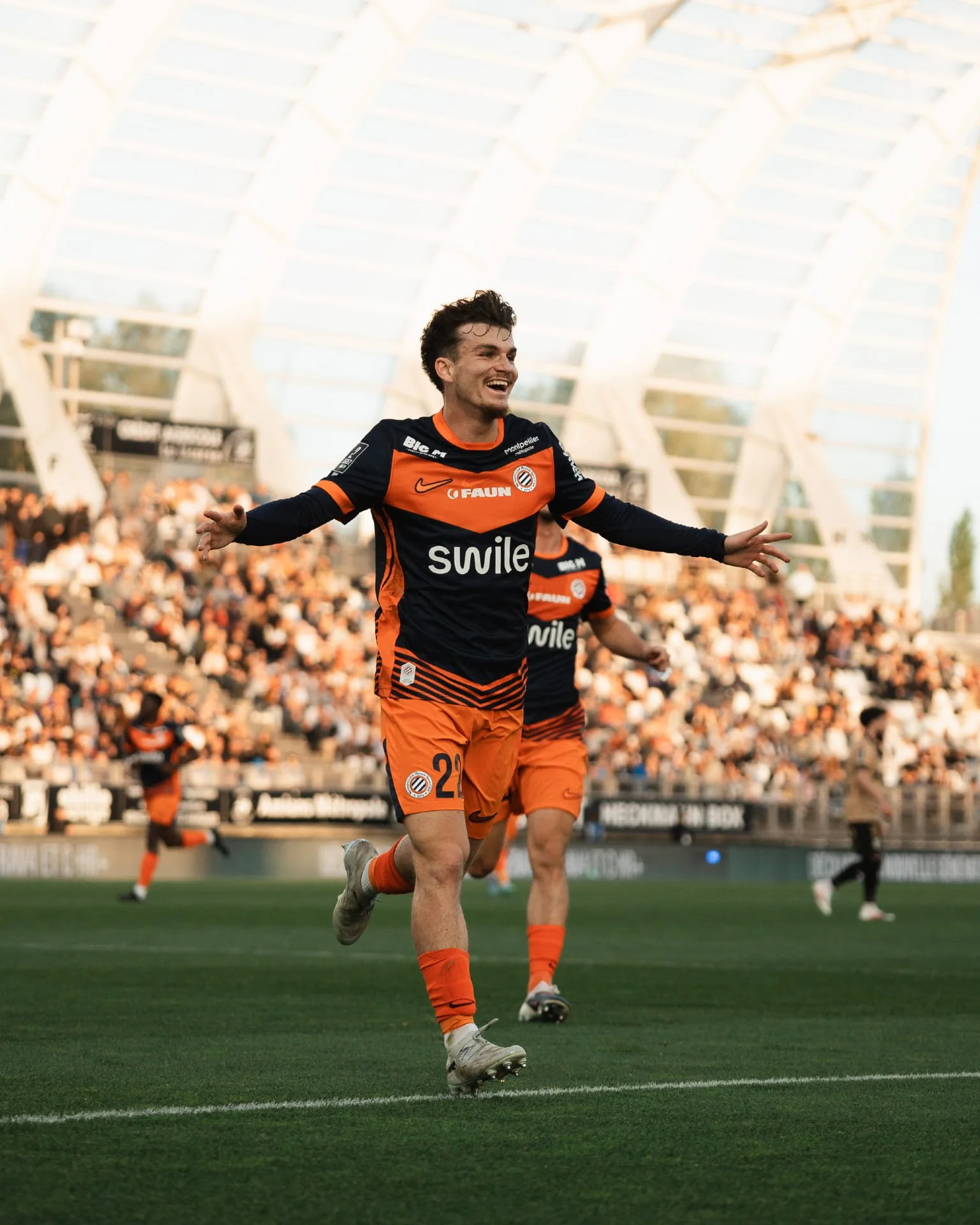 Un joueur de football souriant joue sur un terrain de stade, portant un maillot de l'équipe orange et noire.