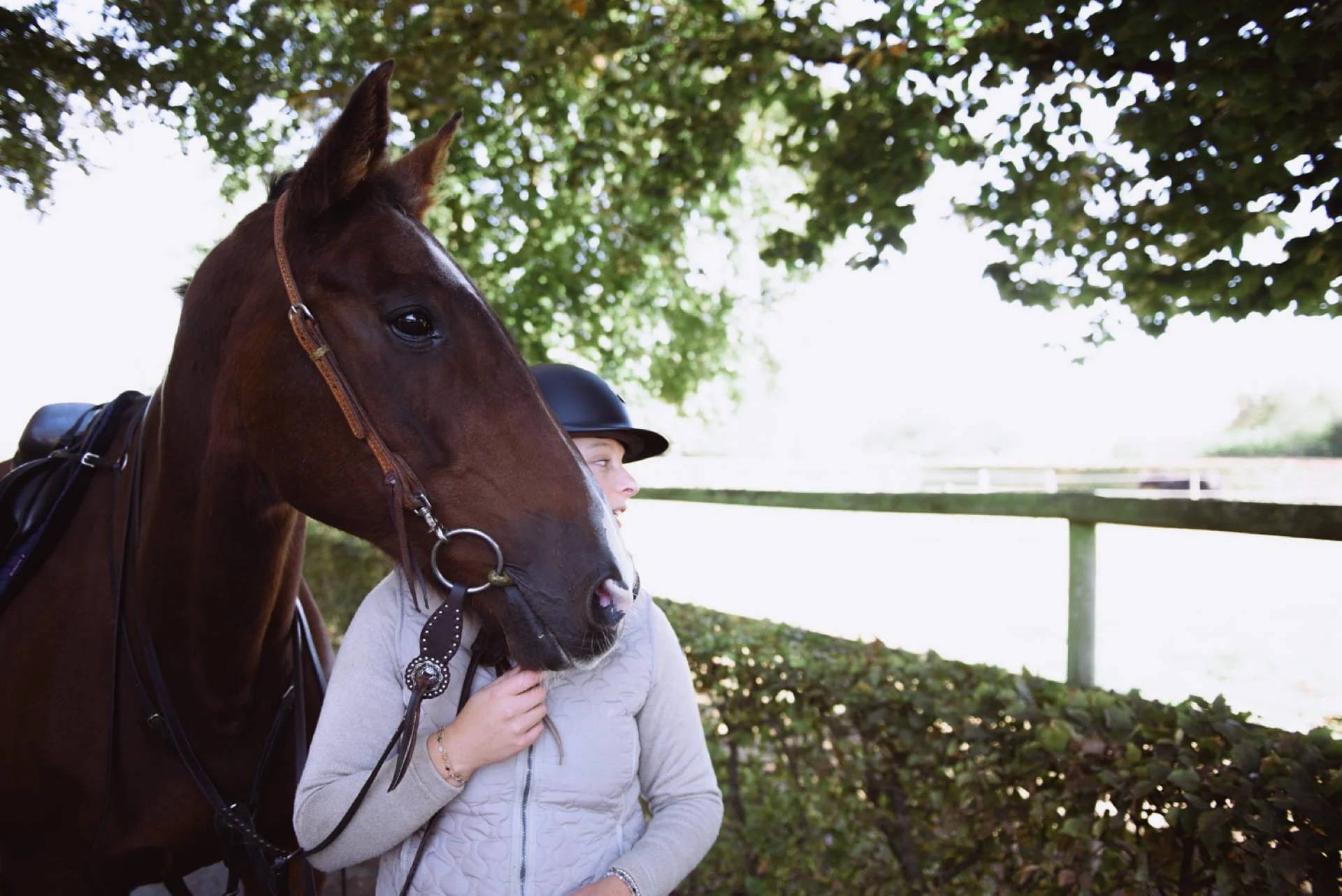 Une femme portant un casque d'équitation et une veste claire est près d'un cheval marron foncé avec une bride en cuir, dans un environnement extérieur ombragé par des arbres. Cauê Foto, photographe Amiens, photographe de mariage et événementiel