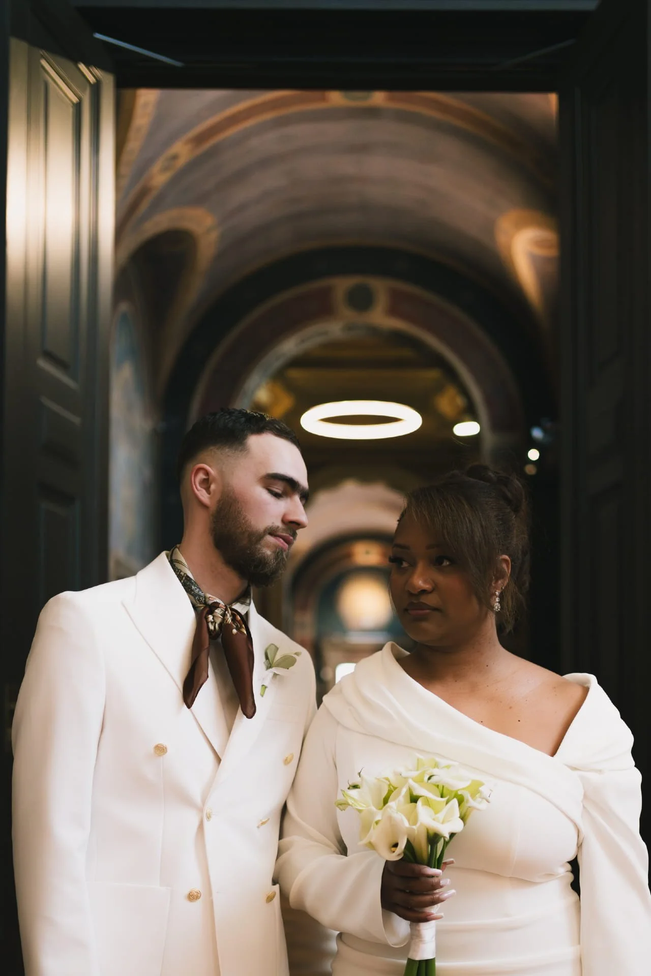 Un couple marié lors d'une cérémonie dans un hall élégant, l'homme porte un costume blanc avec boucles d'oreilles et un foulard, la femme tient un bouquet de fleurs blanches et porte une robe blanche avec des épaules dénudées. Cauê Foto