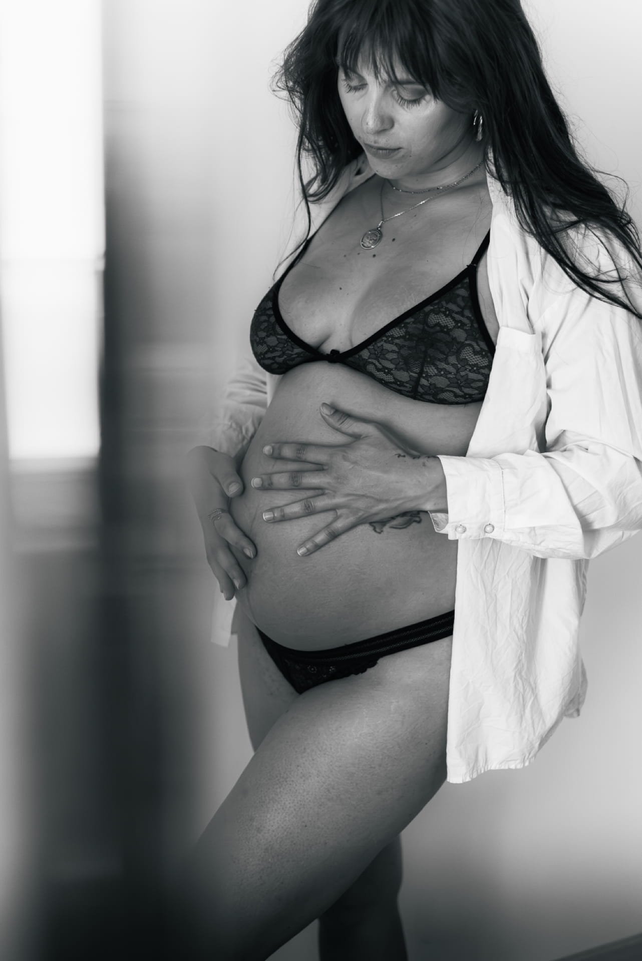 Une femme habillée en lingerie noire, une chemise blanche ouverte, se tient contre un mur, sa main posée sur son ventre. Cauê Foto, photographe Amiens, photographe de mariage et événementiel