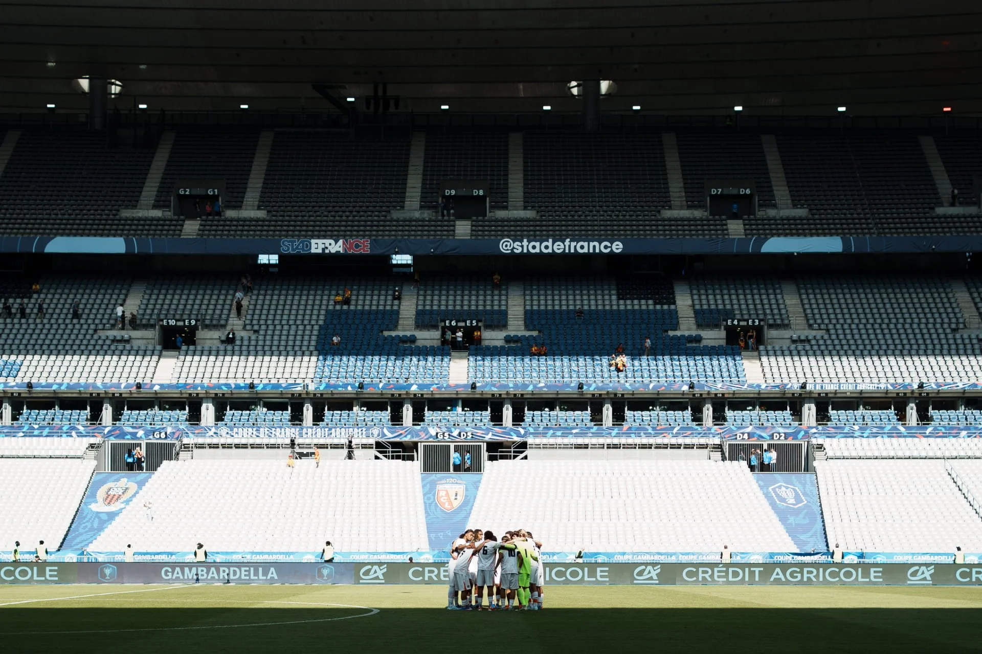Vue intérieure d'un stade de football vide avec des sièges bleus et blancs, et une équipe de football huddle sur le terrain.