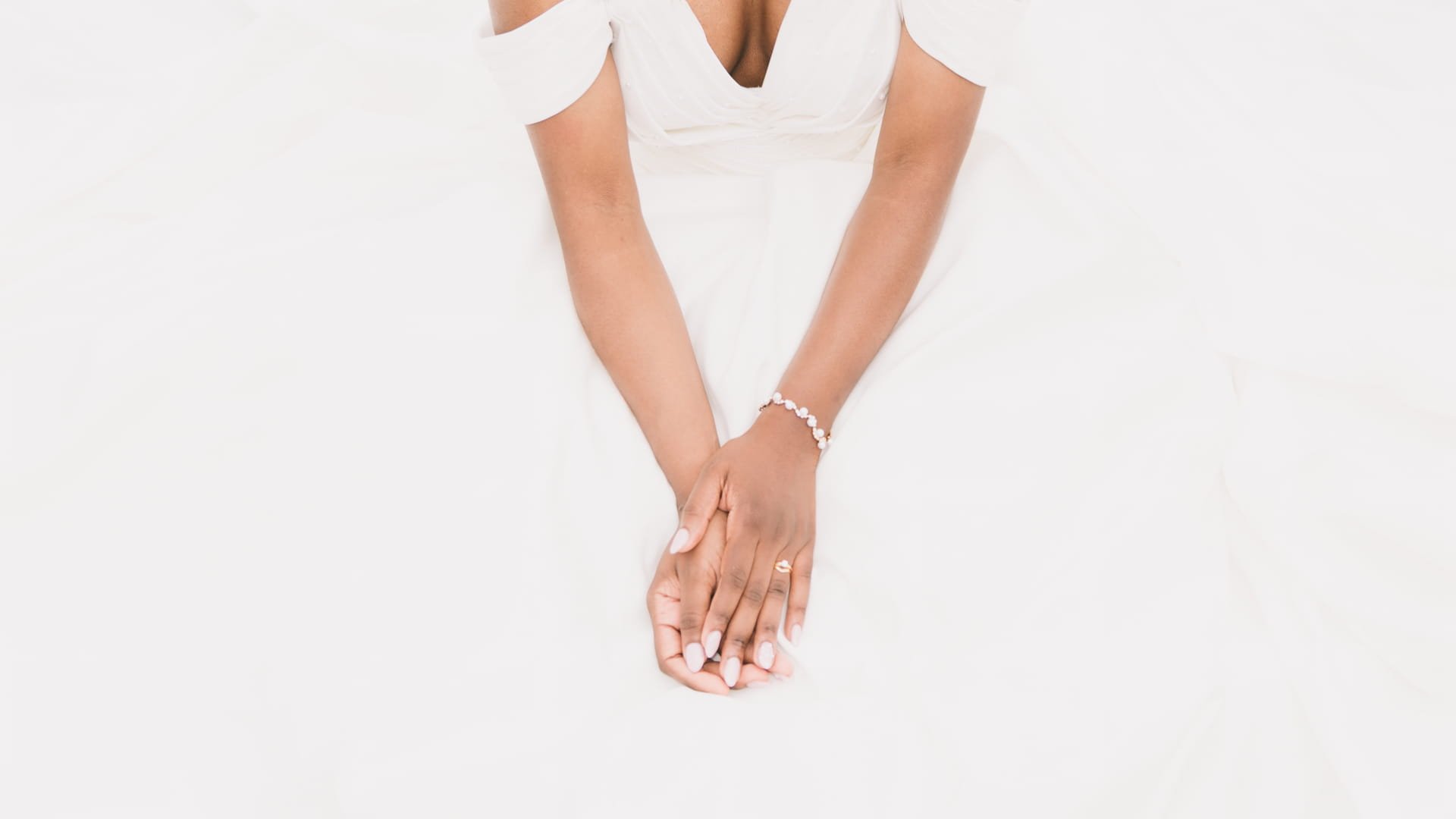 Mains d'une femme avec des ongles manucurés, portant un bracelet et une bague, posé sur une surface blanche, habillée en blanc.