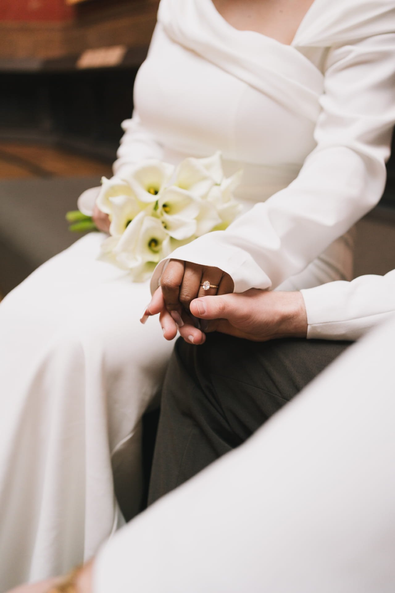 Une personne en robe blanche assise, tenant un bouquet de calla lilies blanches, avec une alliance visible sur son doigt, lors d'une cérémonie.