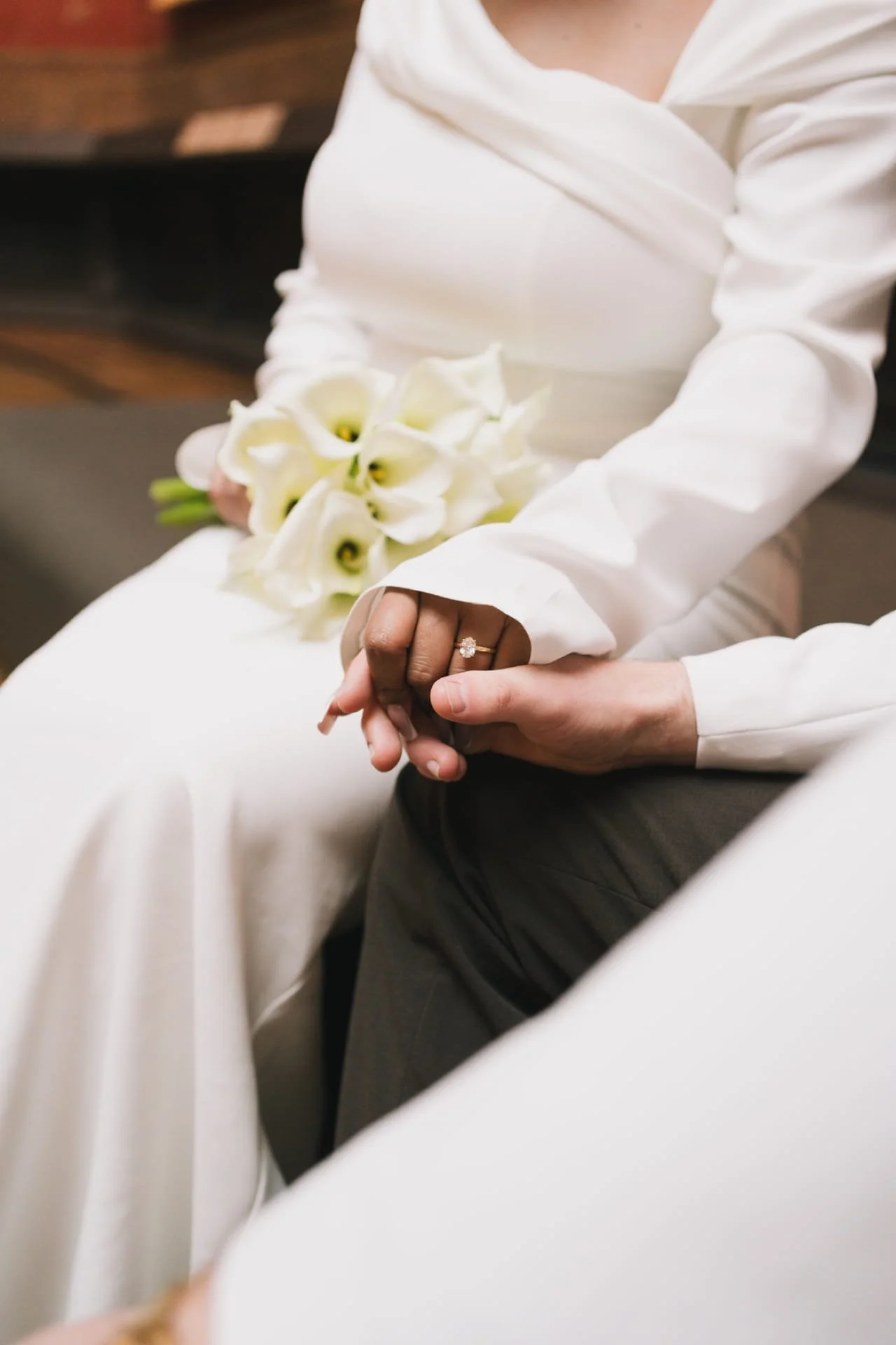 Femme tenant une bouquet de calla lilies, montrant une alliance à l'annulaire, portant une robe blanche avec drapé, assise à côté d'une personne en costumes gris. Cauê Foto, photographe Amiens, photographe de mariage et événementiel