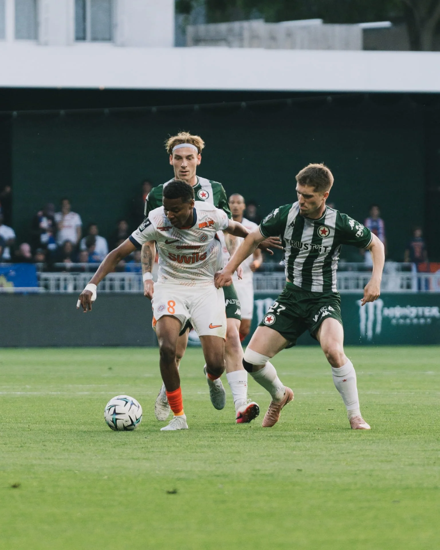 Trois joueurs de football en action lors d'un match, deux en maillots verts et un en blanc, concentrés sur le ballon en pleine partie