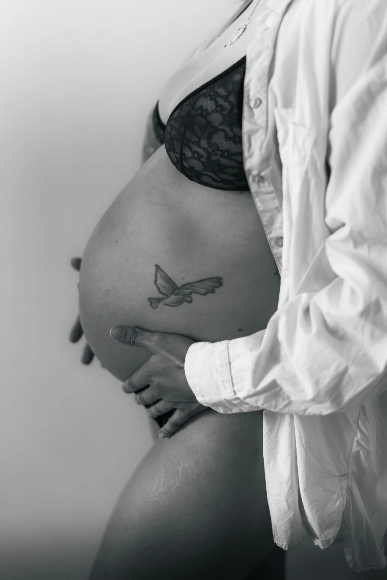 Une femme enceinte porte un soutien-gorge en dentelle noire et une chemise blanche ouverte. Elle tient son ventre arrondi avec ses mains. Une tatouage de papillon est visible sur son abdomen. Cauê Foto, photographe Amiens, photographe de mariage