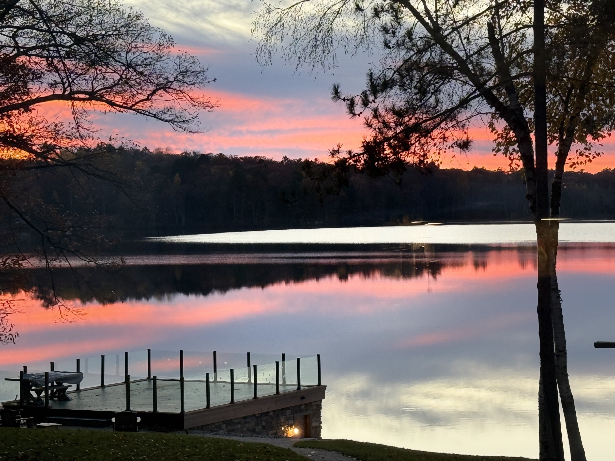 A peaceful lake at sunset with colorful sky, surrounded by trees and with a dock extending into the water.