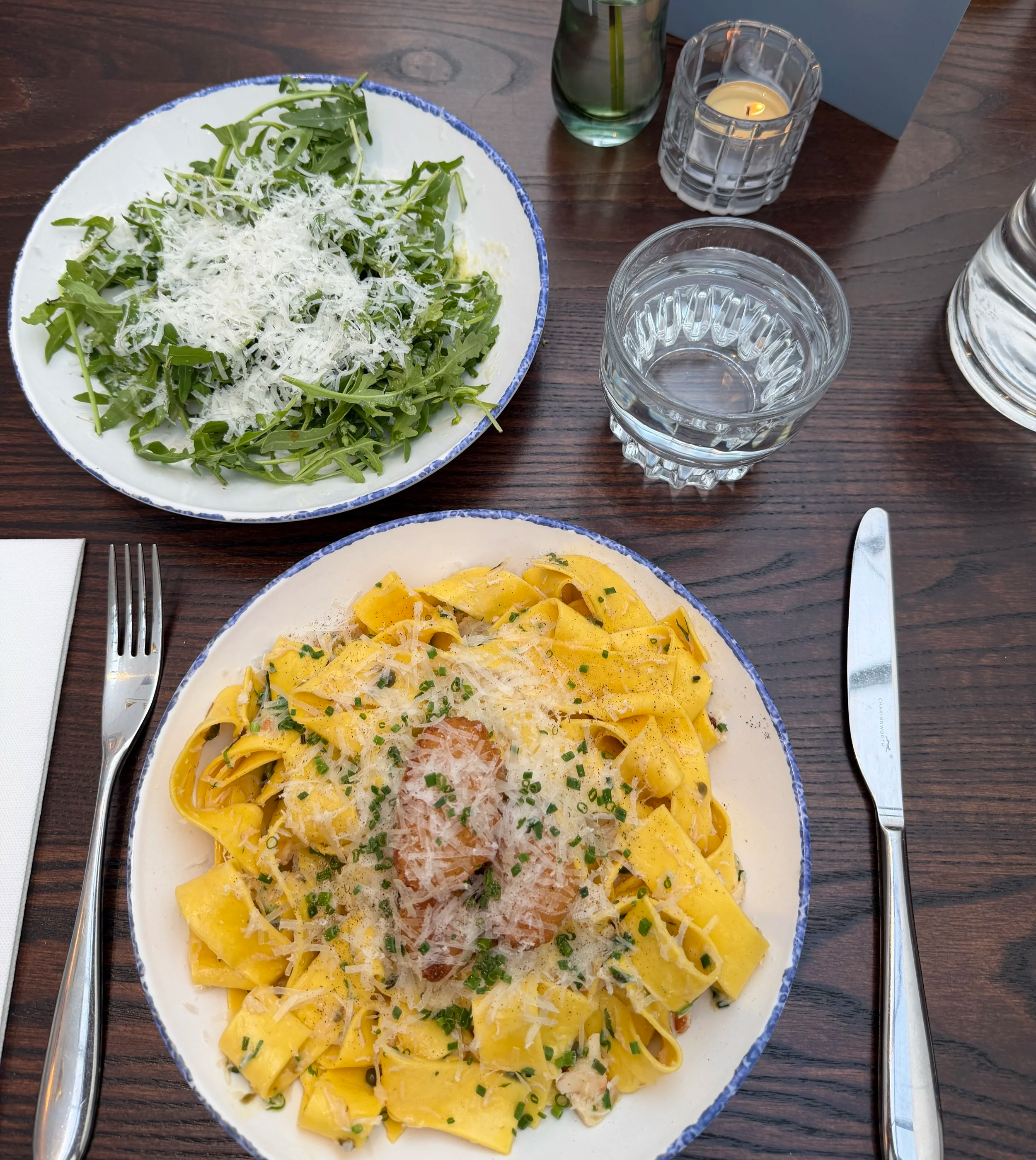 Homemade pappardelle pasta with crab and Whitby scampi on top on a white plate with blue edge and a rocket and parmesan salad in the back also on a white and blue plate.