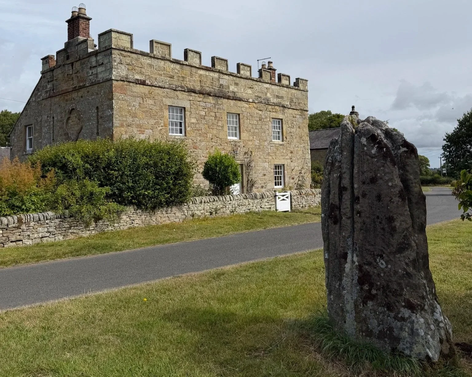 An old stone house in the background with a stone wall and white gate and the Stob stone standing in front.