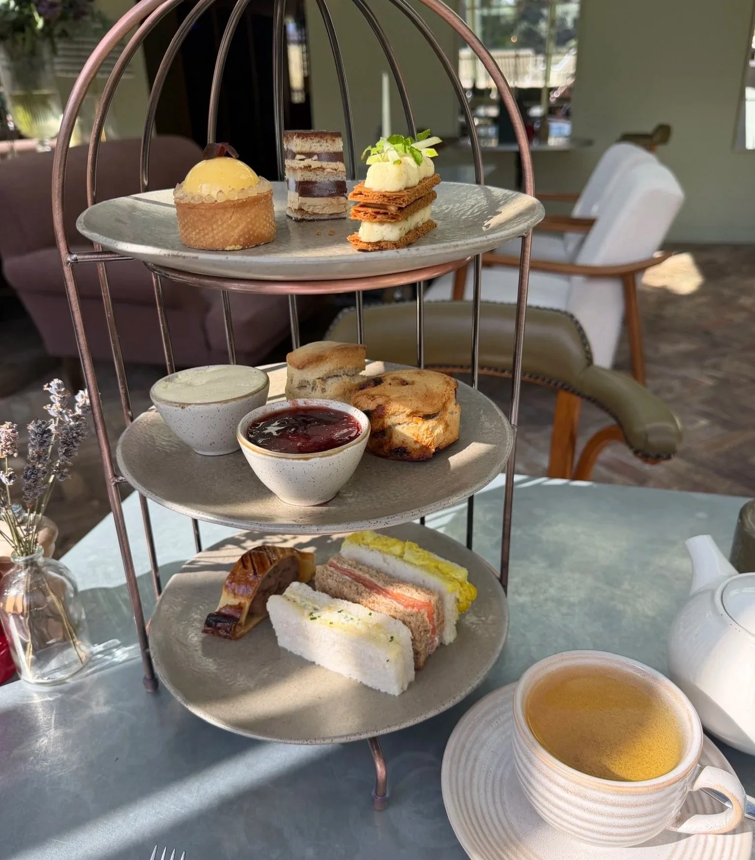 Three tiered afternoon tea with sandwiches and sausage roll, scones with jam and cream and three mini pastries of lemon tart, mille-feiulle and chocolate and espresso cake. A cup of tea sits in the foreground.