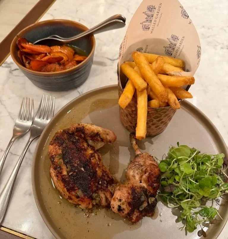 Roast chicken with salad leaves on a round brown plate and fries in a silver cup with walled garden paper and roast carrots in a green dish with a spoon and fork on the lop left corner.