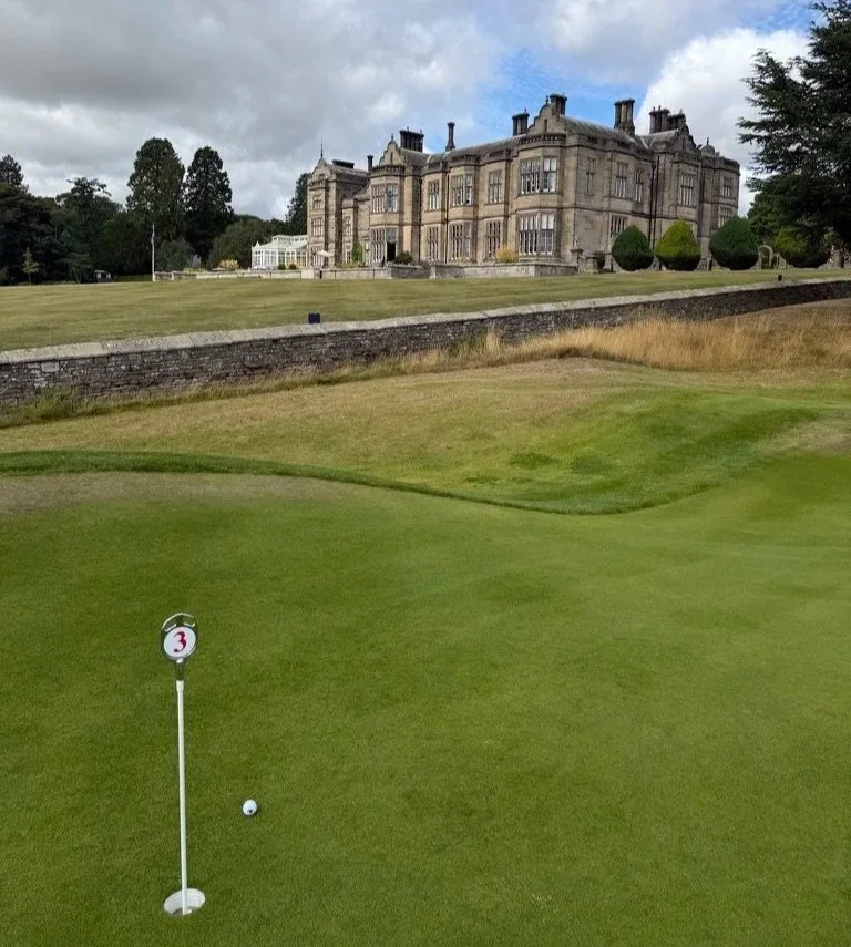 Matfen Hall in the background with one of the holes from the Himalayas course in the foreground with a golf ball beside it.