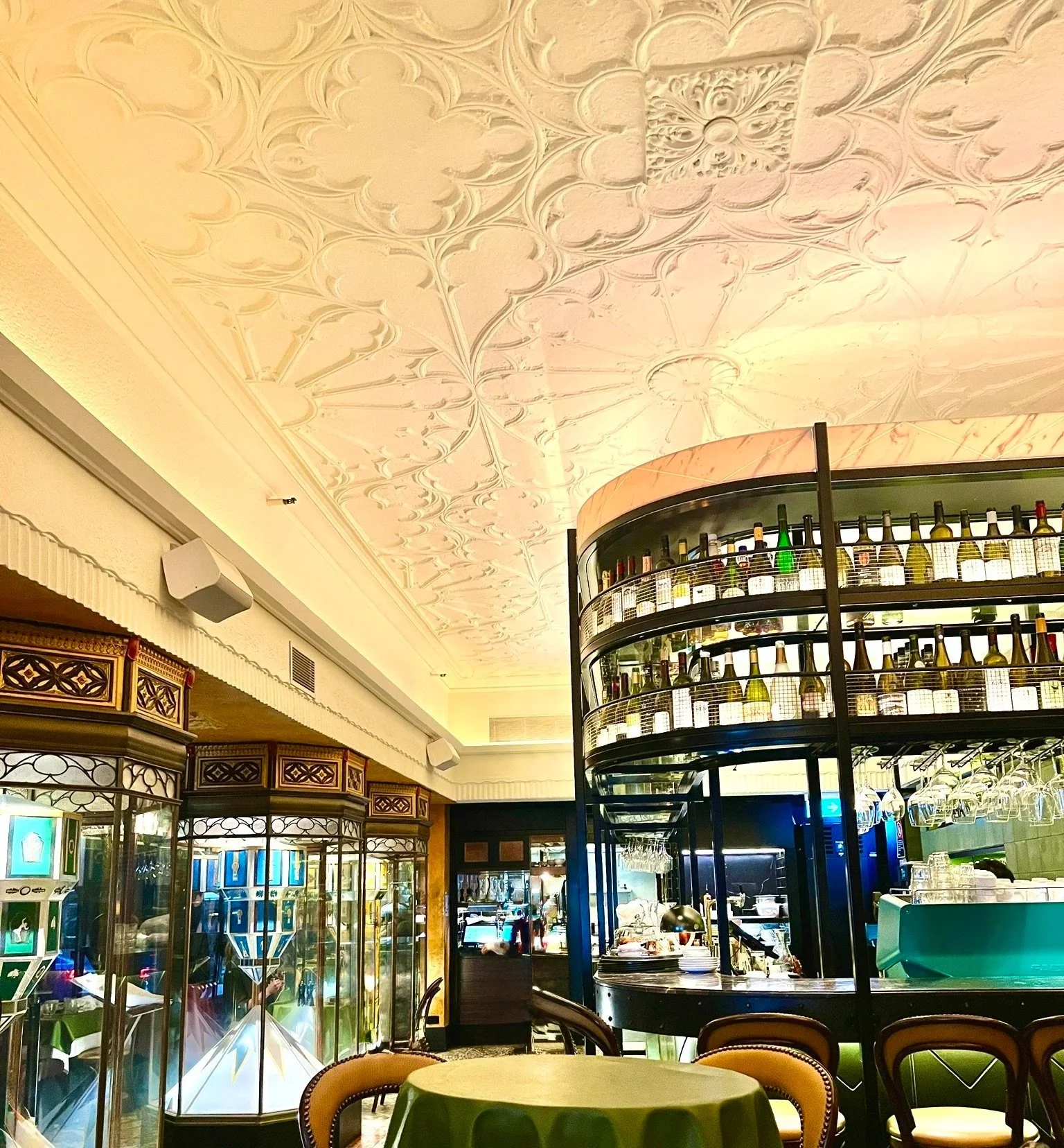 QT Sydney Parlour restaurant art deco patterned ceiling with oval bar with bottles at the top on the right and glass cabinets to the left and chairs and tables at the front.