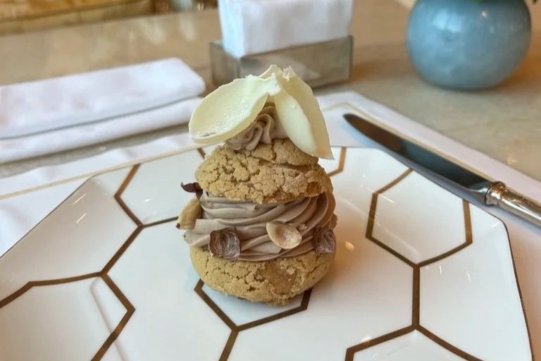 Raffles The Palm Blüthner Hall hazelnut and chocolate choux bun dessert with white chocolate leaf style decoration on white plate with brown geometric lines.